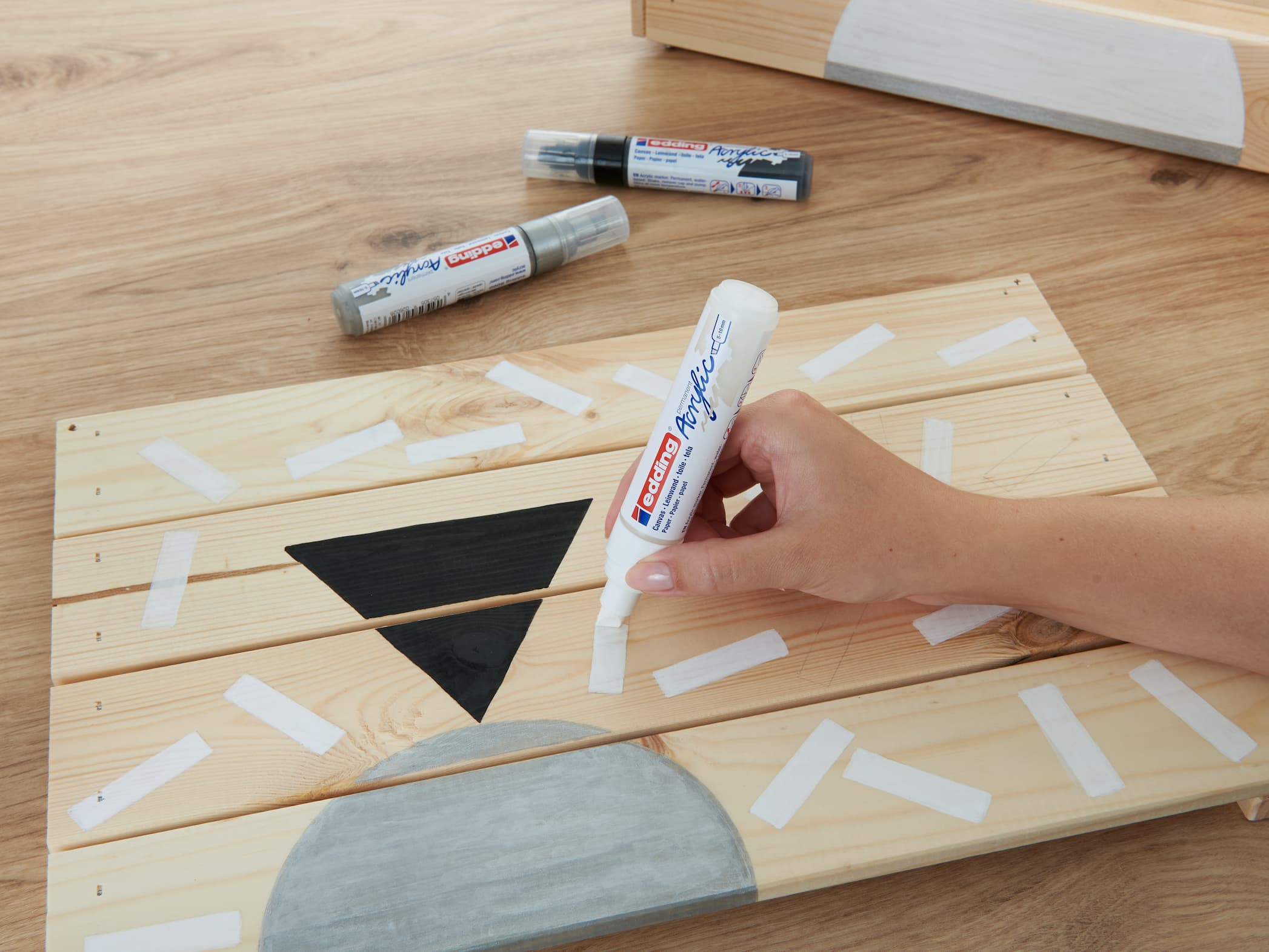 Eine Hand klebt als Vorbereitung für ein Bastel- oder Malprojekt Malerband auf ein Holzbrett, daneben liegen ein paar edding 5000 Acrylmarker breit.