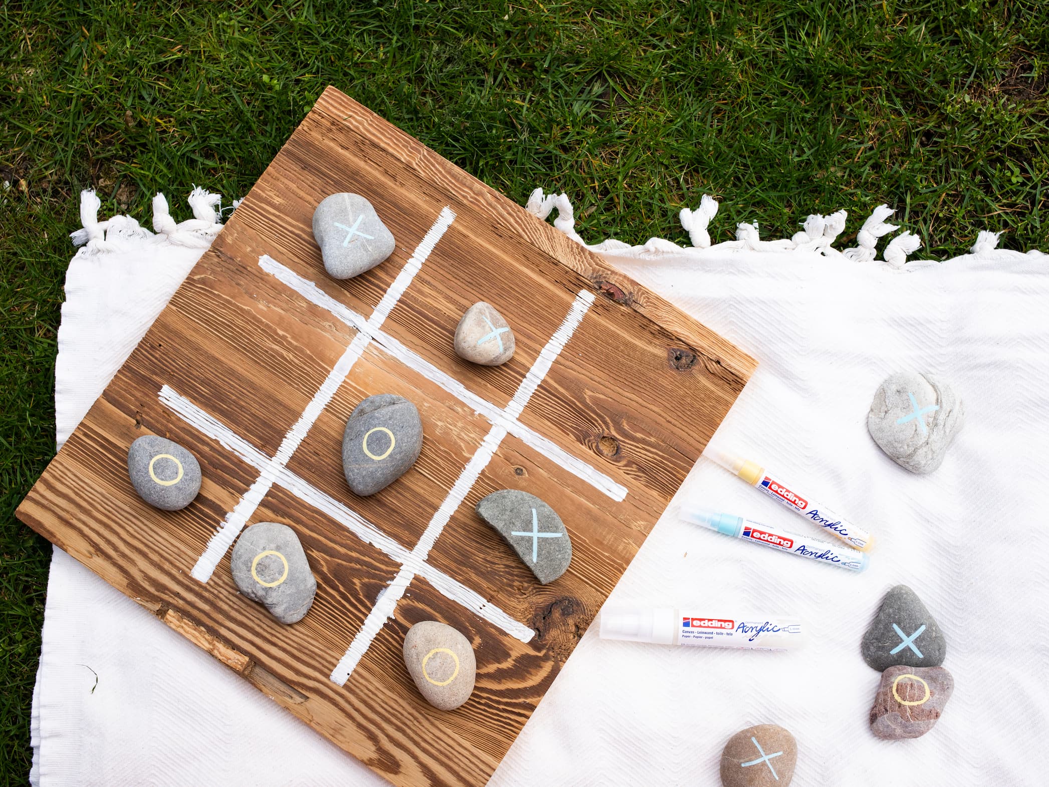 Ein handgefertigtes Tic-Tac-Toe-Brettspiel aus Holz und Steinen, ausgelegt auf einer weißen Decke über Gras, mit Spielmarkierungen, die mit edding 5000 Acrylmarker breit auf die Steine gemalt wurden.