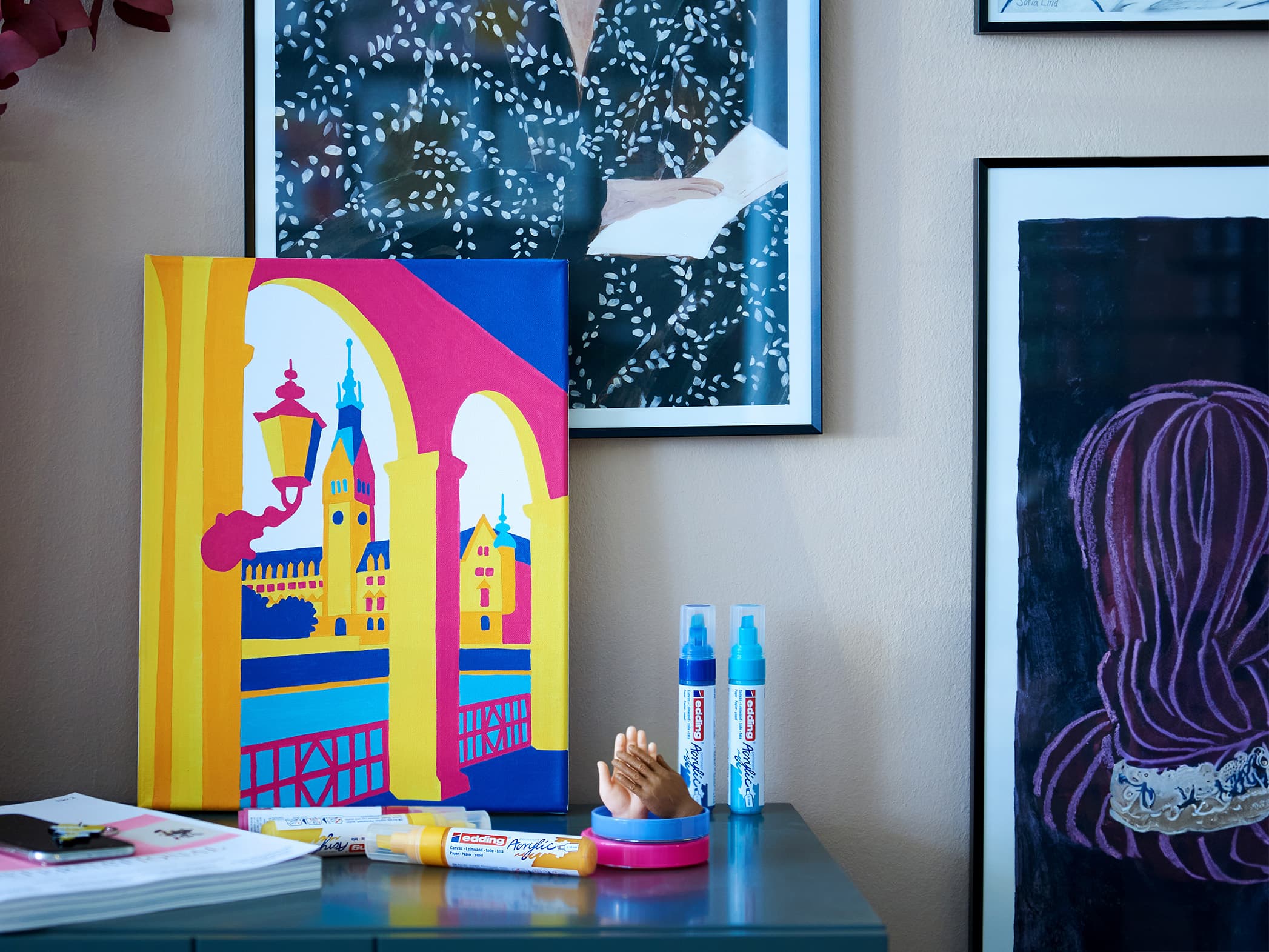 Eine farbenfrohe Ecke mit künstlerischem Flair: An der Wand lehnt ein lebendiges, abstraktes Gemälde architektonischer Merkmale in kräftigen Gelb-, Blau- und Rosatönen, flankiert von einer Sammlung gerahmter Kunstwerke. Ein kleiner edding 5000 Acrylmarker breit.