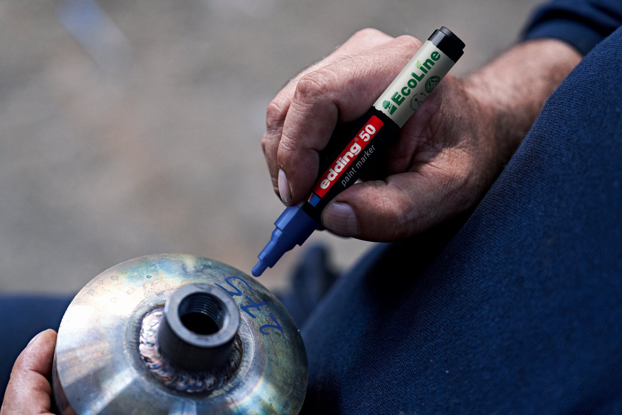 Eine Person verwendet einen edding 50 EcoLine Lackmarker, wasserbasiert, um auf einen Metallgegenstand mit Gewinde und blauen Markierungen zu schreiben. Zu sehen sind lediglich die Hände und ein Teil eines blauen Ärmels. Marke: edding.