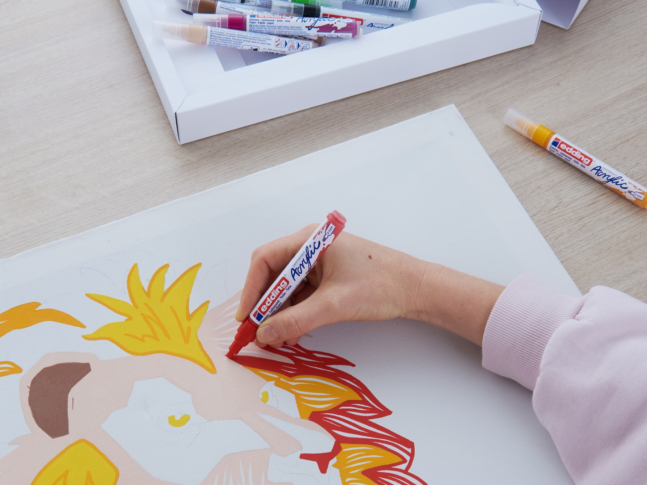 Die Hand eines Künstlers fügt einer Löwenillustration mit einem edding 5100 Acrylmarker-Medium auf einer weißen Leinwand feurig orangefarbene Details hinzu, während Malutensilien auf dem Tisch ausgebreitet sind.