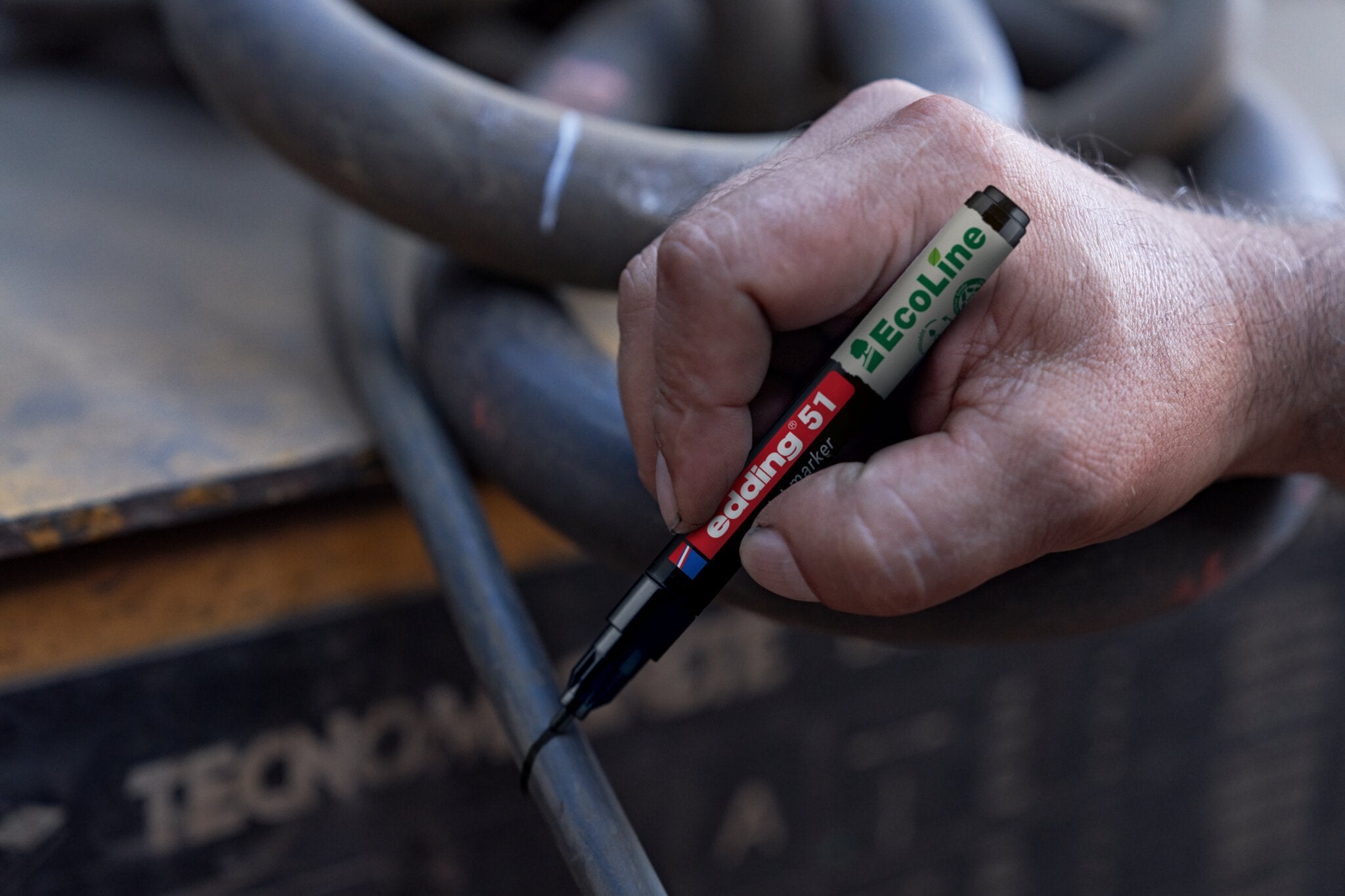 Eine Hand markiert mit einem edding 51 EcoLine Lackmarker, wasserbasiert, eine Linie auf einem schwarzen Kabel, im Hintergrund Industrieanlagen und dicke Kabel – eine nachhaltige Wahl für Profis.