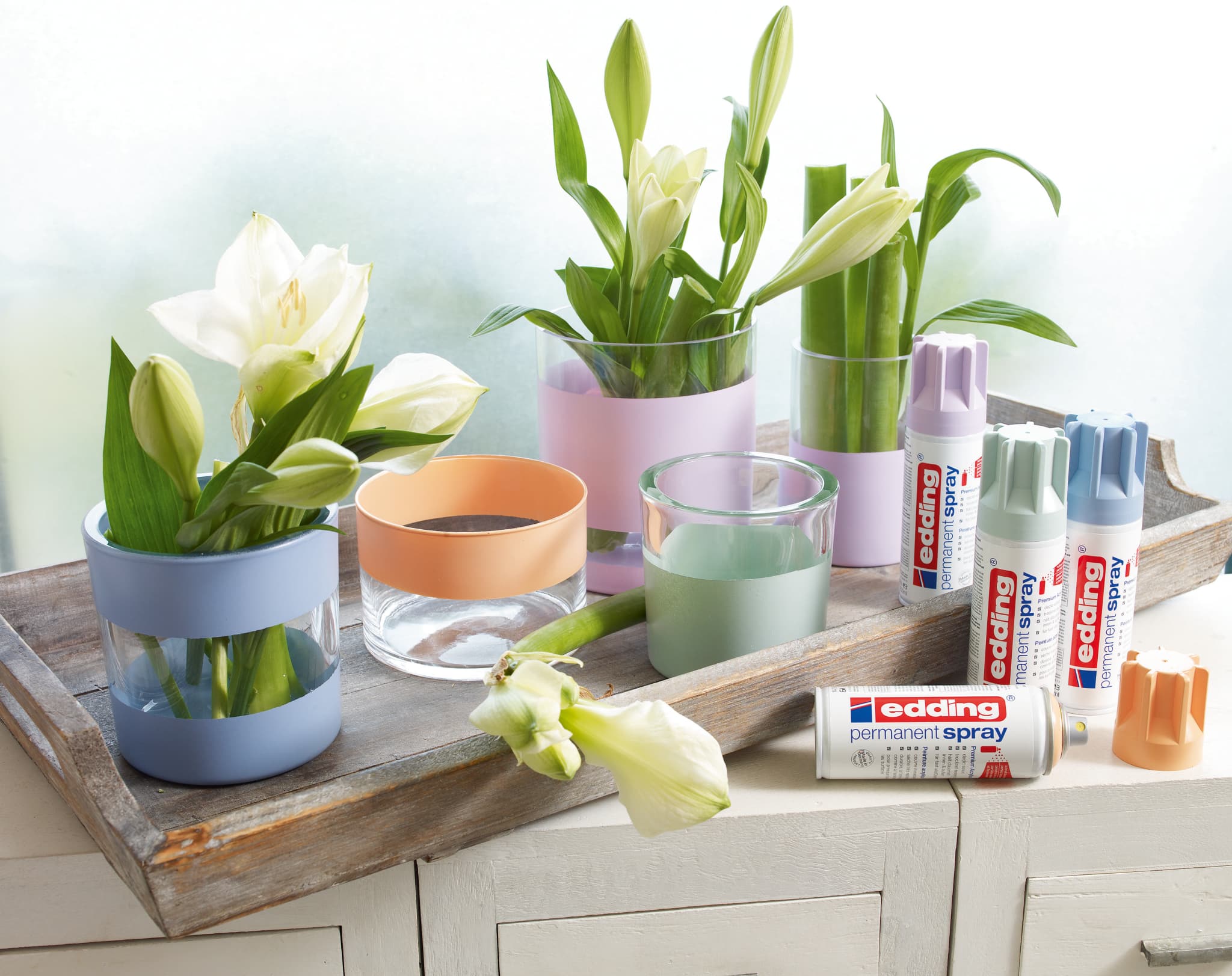 Ein kreatives DIY-Projekt mit wunderschönen weißen Tulpen in frisch gestrichenen Töpfen, begleitet von einer Vielzahl farbenfroher edding 5200 Permanentspray Acryllack Pastell & sanfte Farben-Dosen auf einem rustikalen Holztisch, bereit für eine Bastelsession