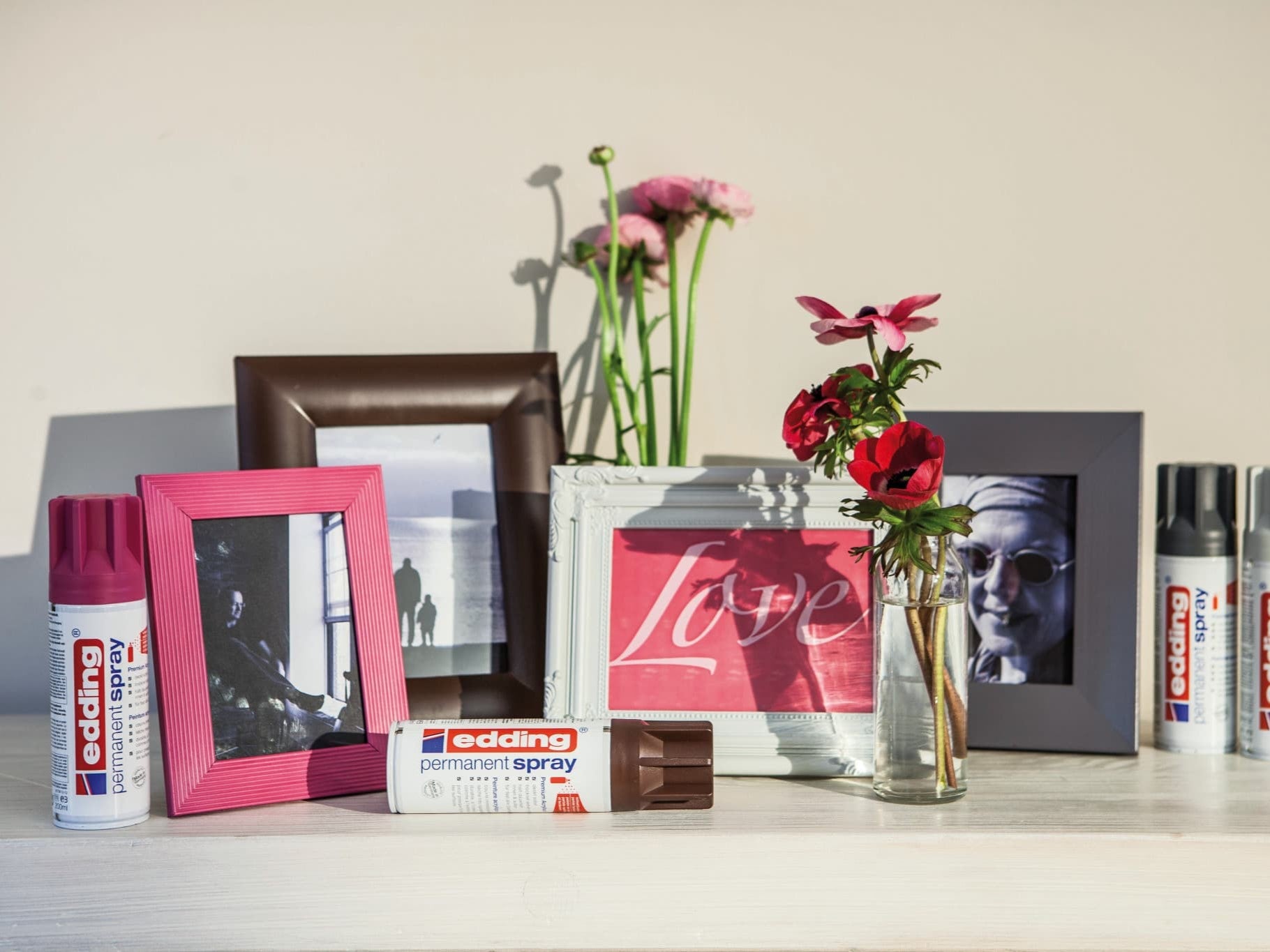Ein kreativer Arbeitsplatz mit edding 5200 Permanentspray Acryllack Neon & Kräftige Farben-Sprühdosen, lebendigen Bilderrahmen, einer Glasvase mit frischen Blumen und inspirierenden gerahmten Kunstwerken vor einem neutralen Hintergrund.
