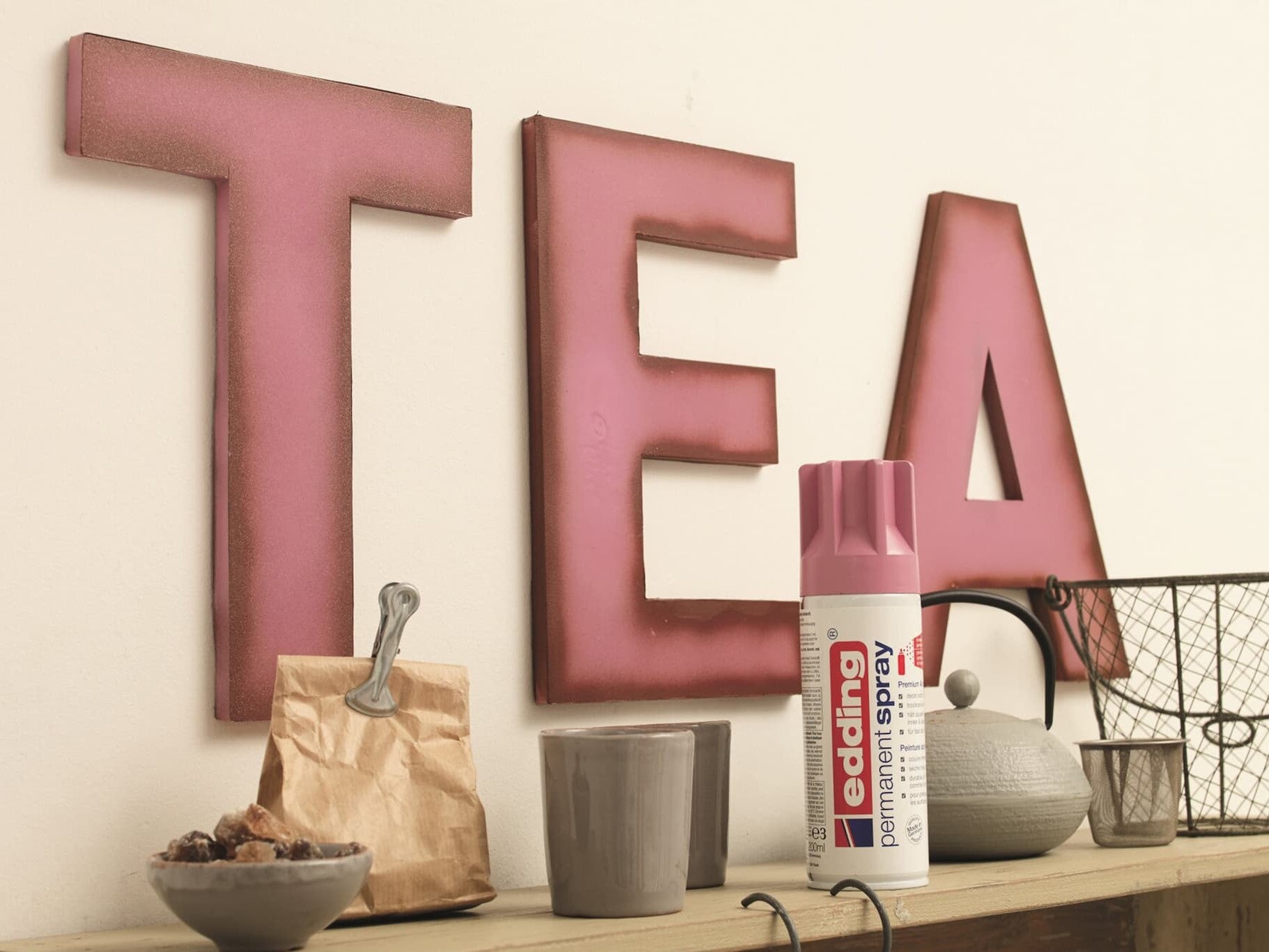 Große Buchstaben mit der Aufschrift „Tee“ auf einem Regal, begleitet von einer kleinen braunen Papiertüte, einer Metallschaufel, einem grauen Becher, einer Dose edding 5200 Permanentspray Acryllack Pastell & sanfte Farben und Dekorationsartikeln in Past.