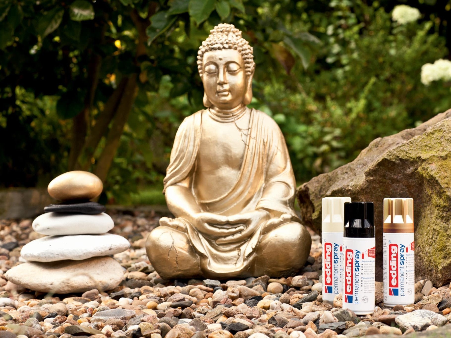Eine ruhige Gartenkulisse mit einer goldenen Buddhastatue, die neben einem ausbalancierten Steinstapel meditiert, flankiert von drei Tuben edding-Kleber auf einem Kieselsteinbett, das mit edding 5200 Permanentspray Metallic Acryllack beschichtet ist.