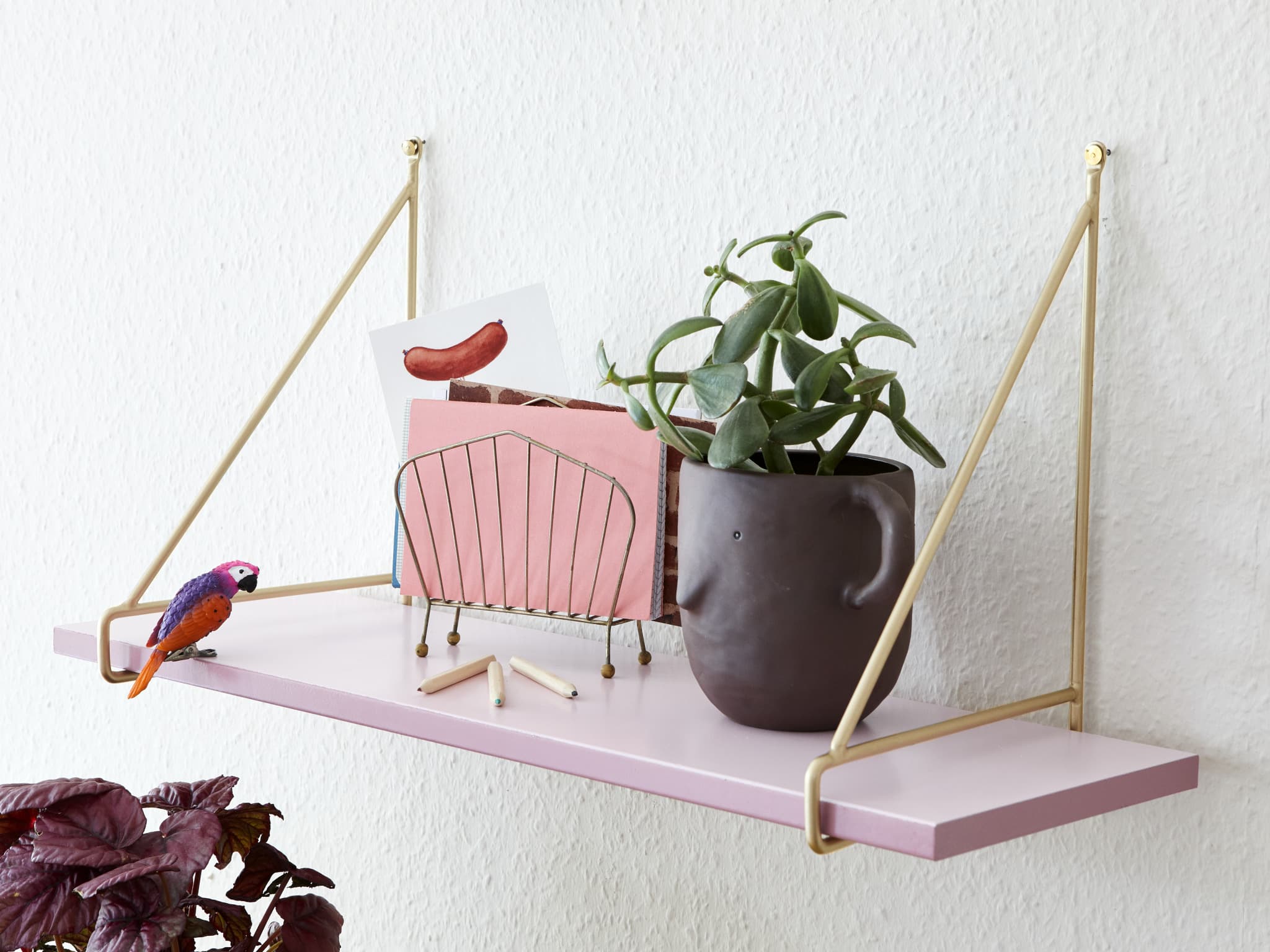 Ein stilvoll dekoriertes Wandregal mit einer kleinen Pflanze in einem braunen Topf, einem gerahmten abstrakten Kunstwerk und einer farbenfrohen Vogelfigur, alles mit minimalistischer Eleganz verbunden und durch edding 5200 Permanentspray Metallic Acryllack aufgewertet.
