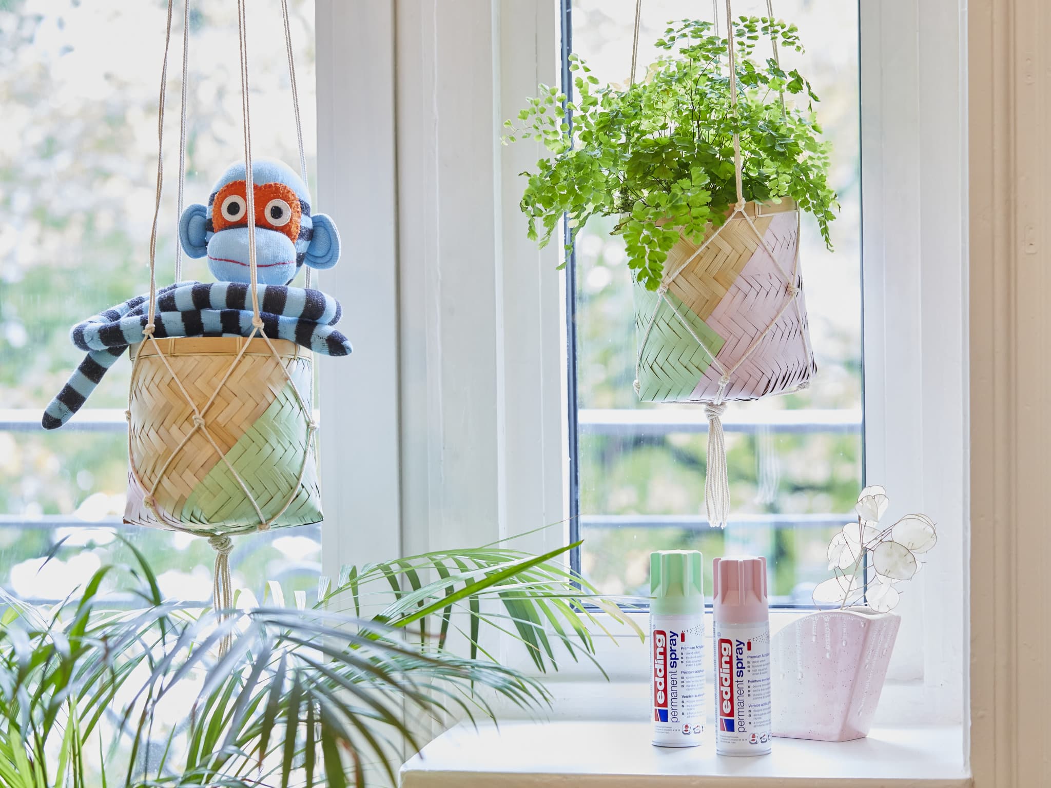 Ein farbenfrohes Sockenaffenspielzeug hängt neben einer fröhlichen Topfpflanze am Fenster, und auf der Fensterbank stehen Künstlerbedarfsartikel wie edding 5200 Permanentspray Acryllack Pastell & sanfte Farben, bereit für einen Tag voller Kreativität.