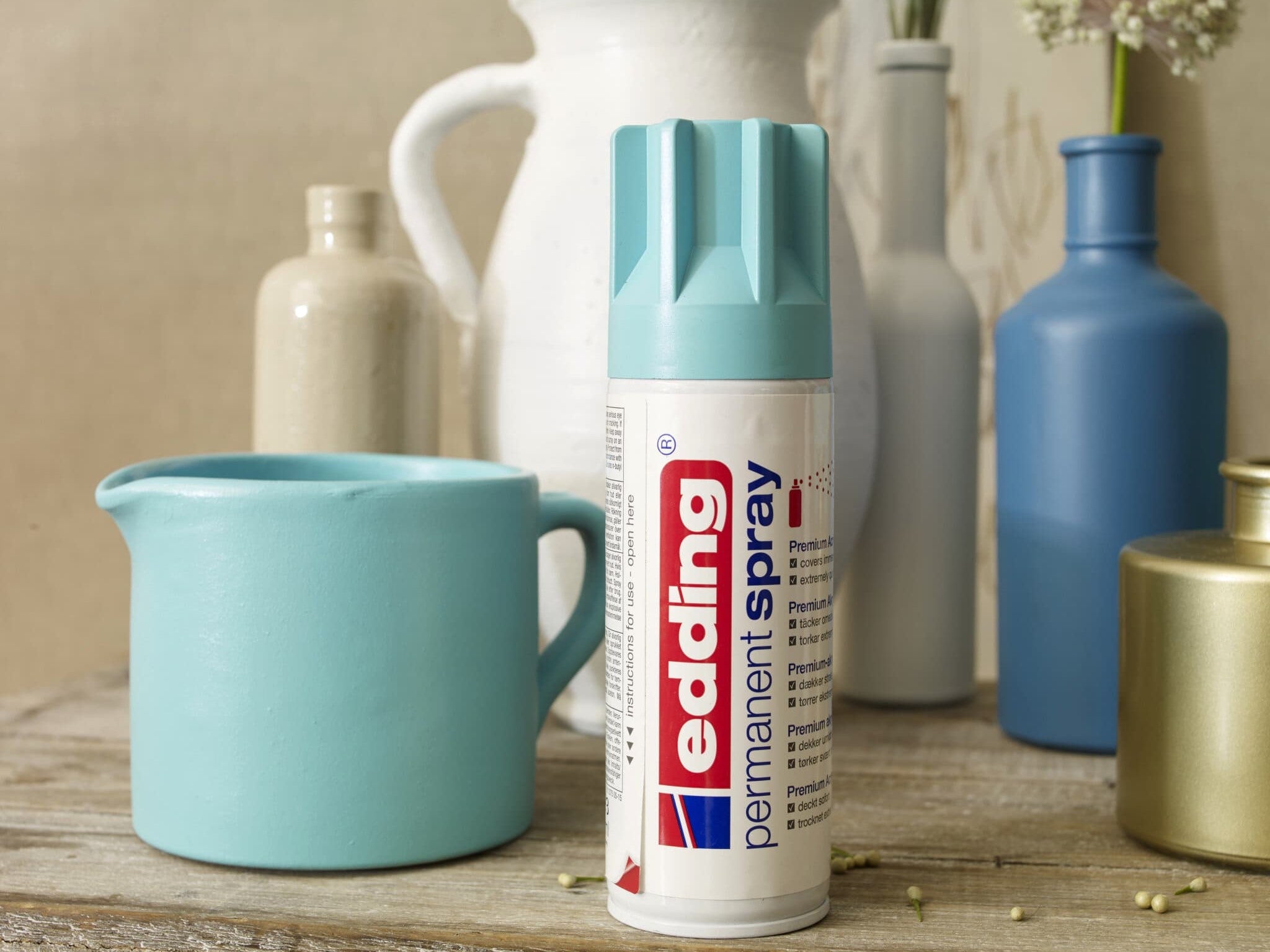 Eine Dose edding 5200 Permanentspray Acryllack Pastell & sanfte Farben-Sprühfarbe ist inmitten einer Sammlung verschiedenartig geformter und farbiger Vasen in Pastelltönen auf einer rustikalen Holzoberfläche ausgestellt und erinnert an ein Heimwerkerprojekt oder ein Heimwerken.