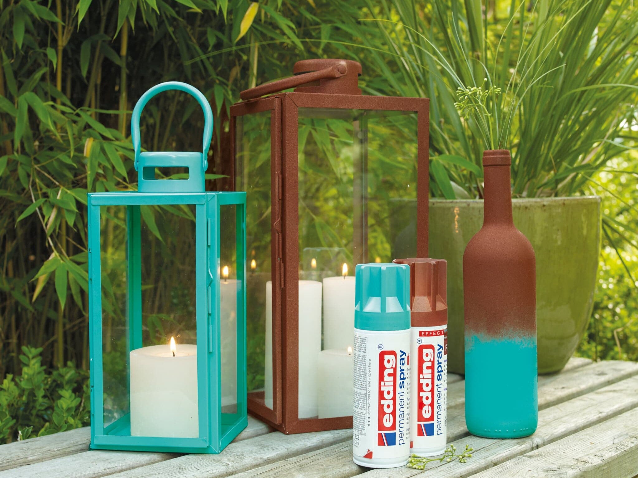 Eine ruhige Außenkulisse mit zwei dekorativen Laternen, eine davon mit einer Kerze beleuchtet, neben einer kreativ mit edding 5200 Permanentspray Effekt Acryllack besprühten Flasche, alles auf einem Holztisch arrangiert.