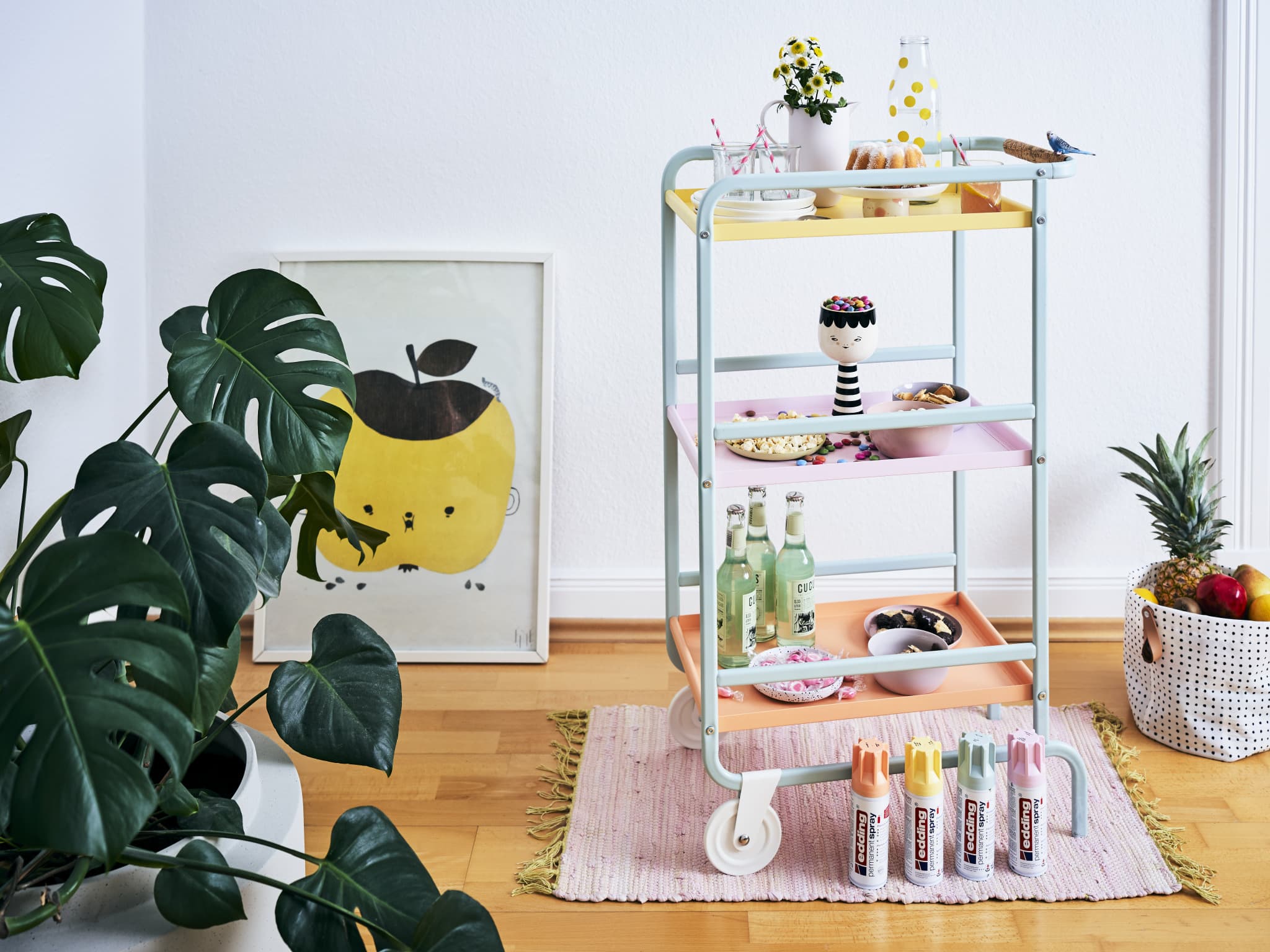 Ein fröhlicher Servierwagen in Pastellfarben mit Snacks, Getränken und Künstlerbedarf, ergänzt durch farbenfrohe Zimmerpflanzen und verspielte Kunst in einer gemütlichen Ecke. Das gesamte Set besteht aus einem edding 5200 Permanentspray Acryllack Pastell & sanfte Farben.