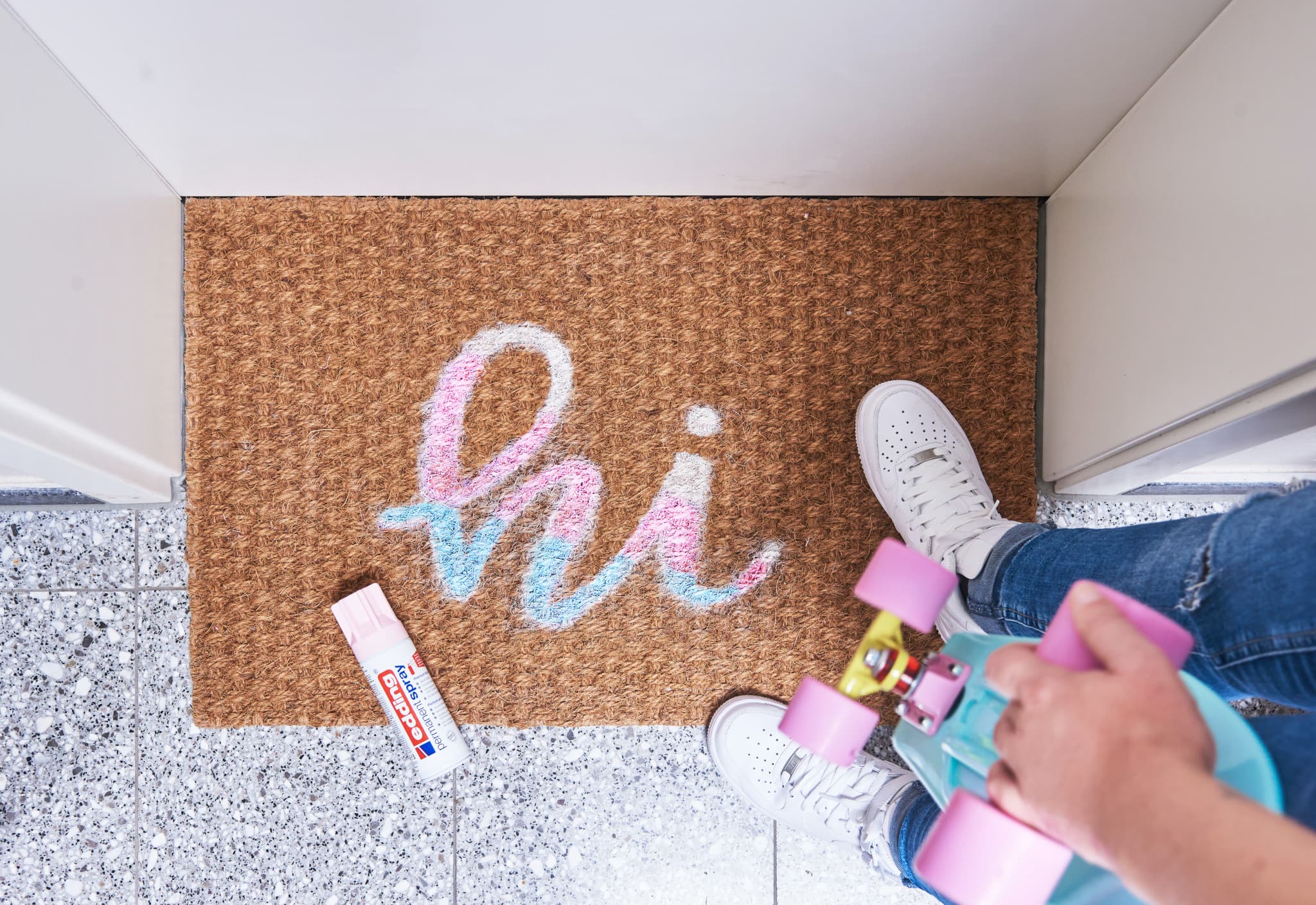 Eine Person steht auf einer Fußmatte mit der Aufschrift „Hi“ in edding 5200 Permanentspray Acryllack Pastell & sanfte Farben und hält ein Skateboard und eine Tube Creme in der Hand, was eine zwanglose und freundliche Begrüßung vor der Haustür suggeriert.
