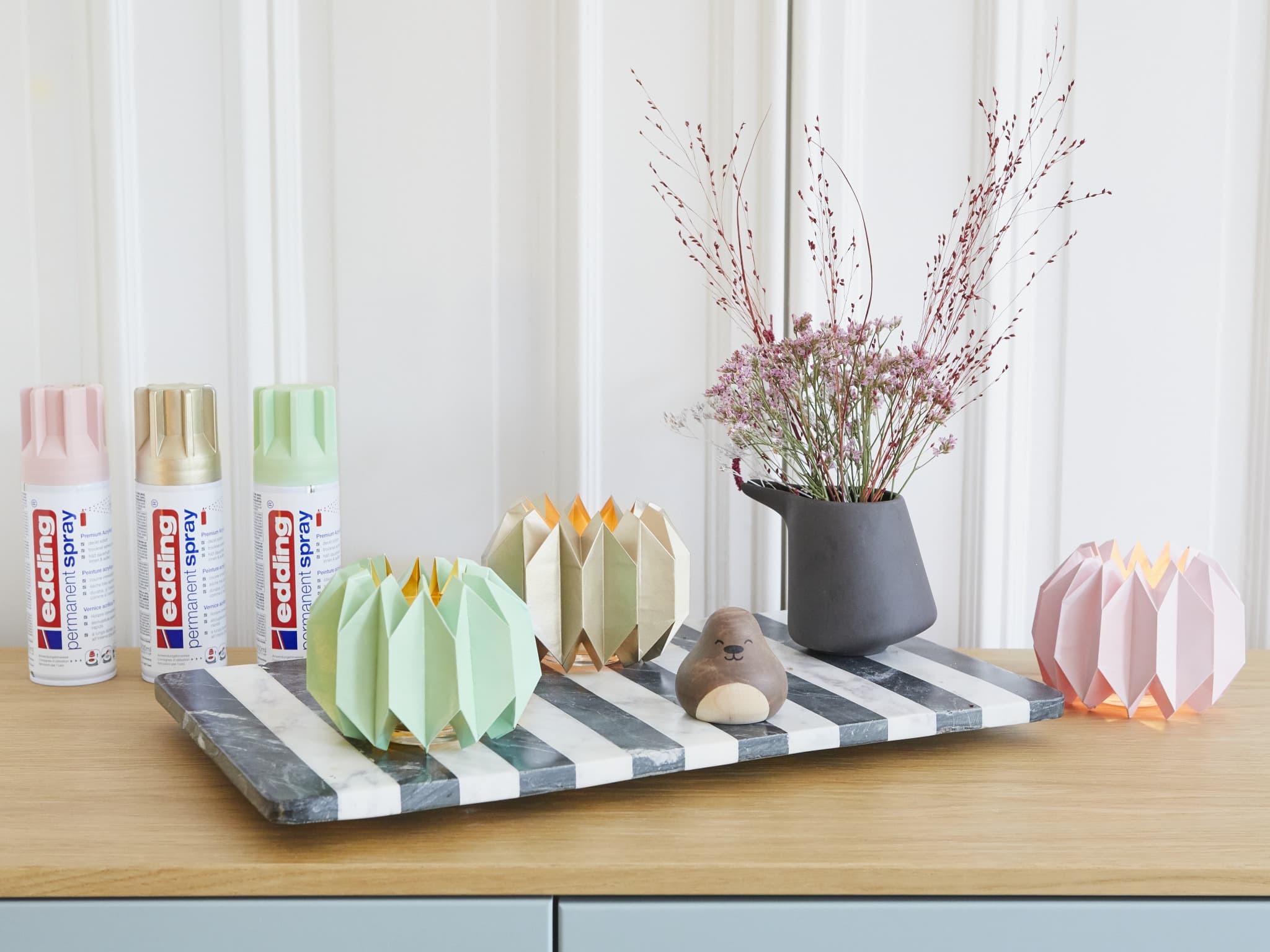 Ein moderner und kreativer Arbeitsplatz, an dem eine Sammlung pastellfarbener edding 5200 Permanentspray Acryllack-Dosen, eine Auswahl farbenfroher gefalteter Papierdekore und eine Vase mit zarten rosa Blumen präsentiert werden, alles hübsch arrangiert.