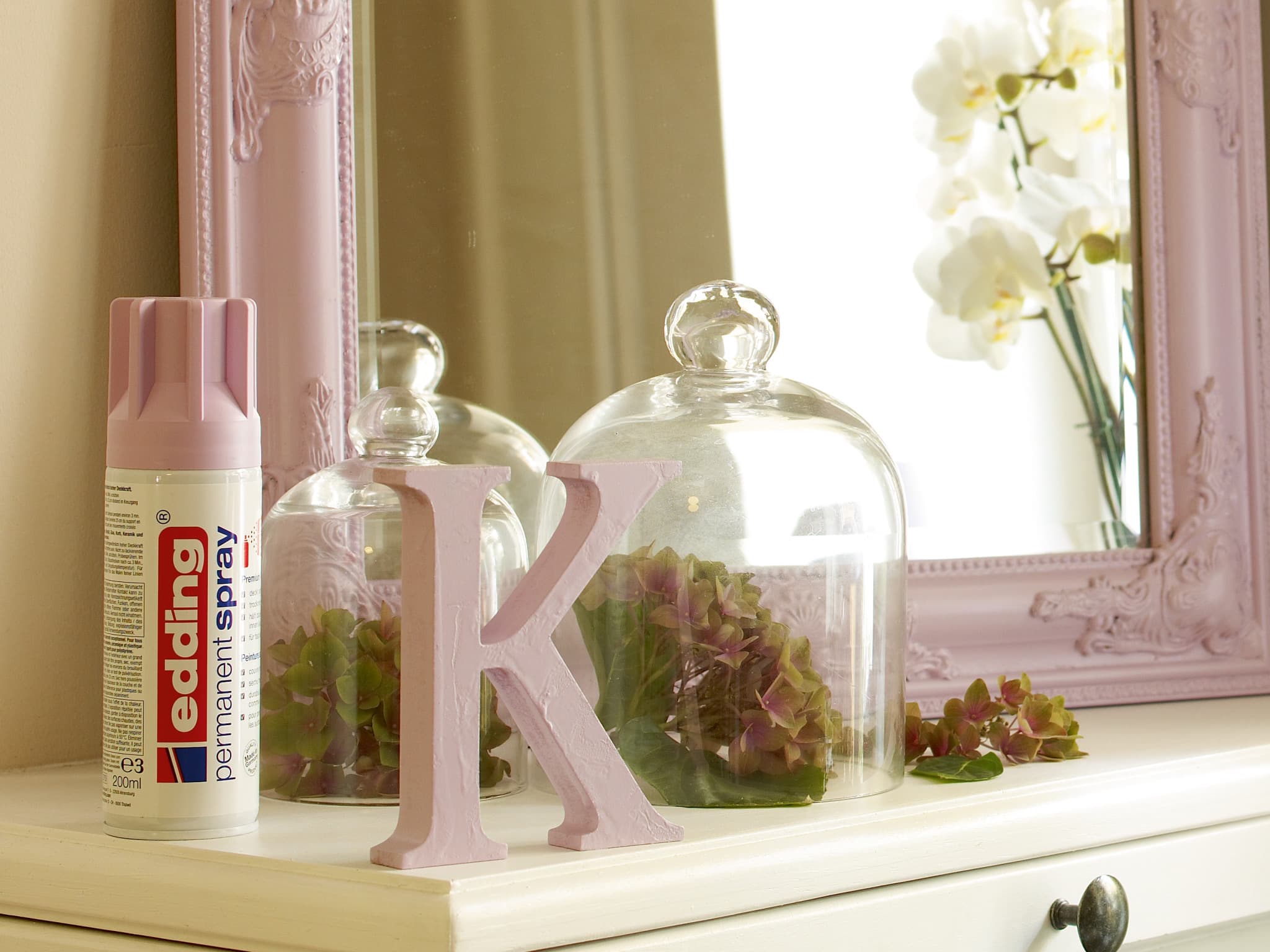 Eine gemütliche Schminkecke mit femininem Touch, mit einem edding 5200 Permanentspray Acryllack Pastell & sanfte Farben, einem dekorativen Buchstaben „k“, zwei Glasglocken mit Blumenarrangements und einem passenden verzierten Spiegel, der