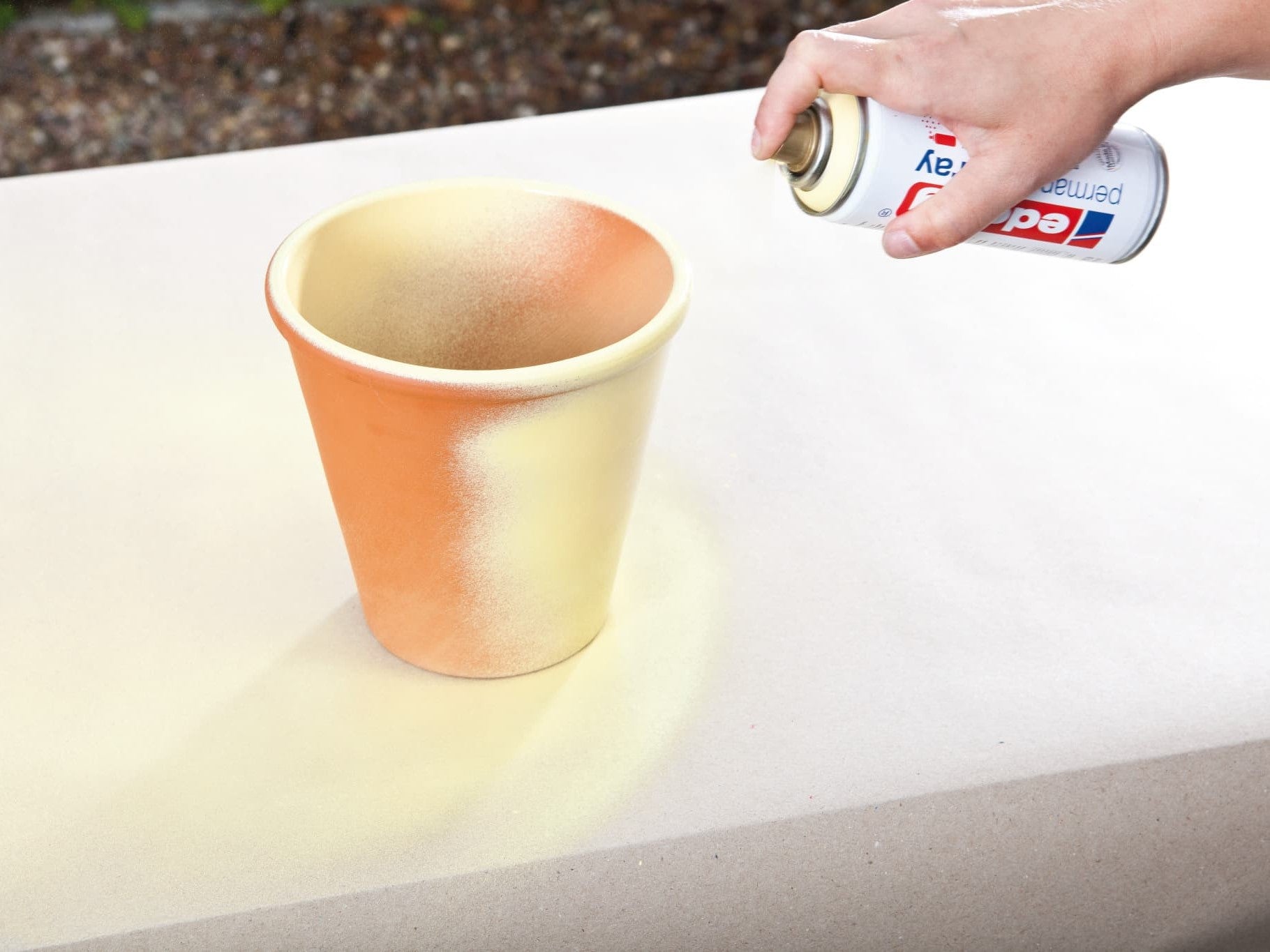 Eine Hand sprüht mit edding 5200 Permanentspray Acryllack Pastell & sanfte Farben Goldfarbe auf einen Terrakotta-Blumentopf und verleiht ihm so eine kreative und persönliche Note.