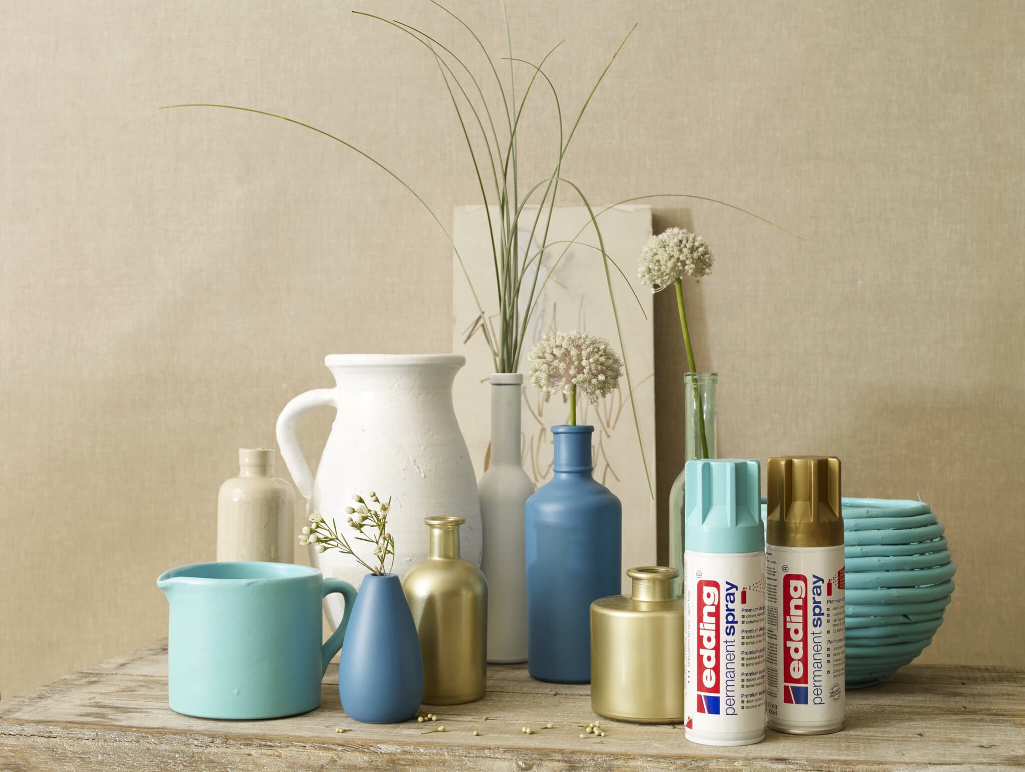 Ein ruhiges Stillleben mit einer Sammlung von Keramikvasen in neutralen Tönen, kombiniert mit Dosen edding 5200 Permanentspray Metallic Acryllack auf einem Tisch, vermittelt den Eindruck kreativen Potenzials in der Inneneinrichtung.