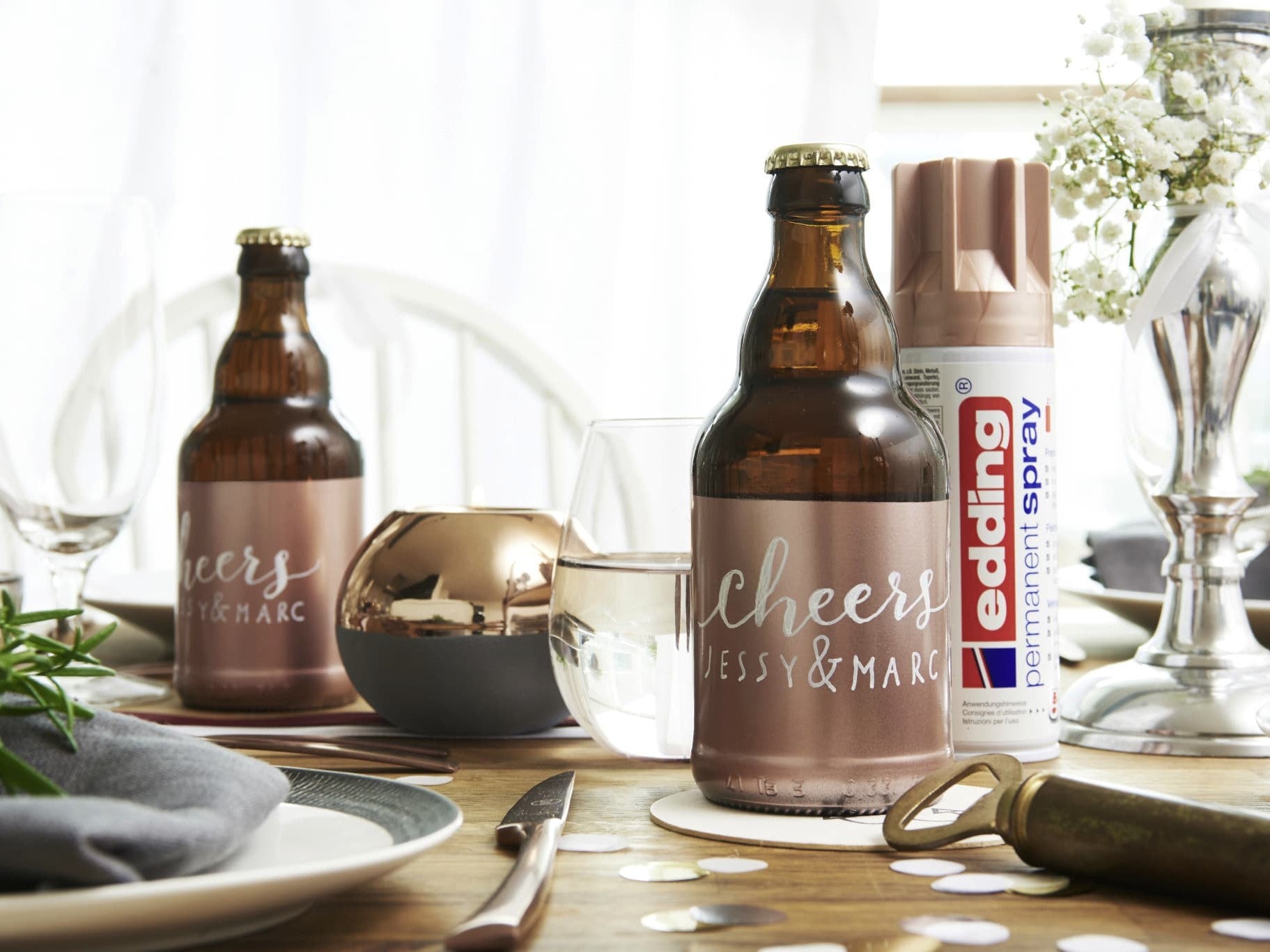 Ein geschmackvoll gedeckter Esstisch mit personalisierten Bierflaschen mit der Aufschrift „Cheers by Marc & Jessy“, begleitet von elegantem Geschirr, Besteck und einem kleinen Blumenarrangement mit einem edding 5200 Permanentspray Metallic Acryllack.