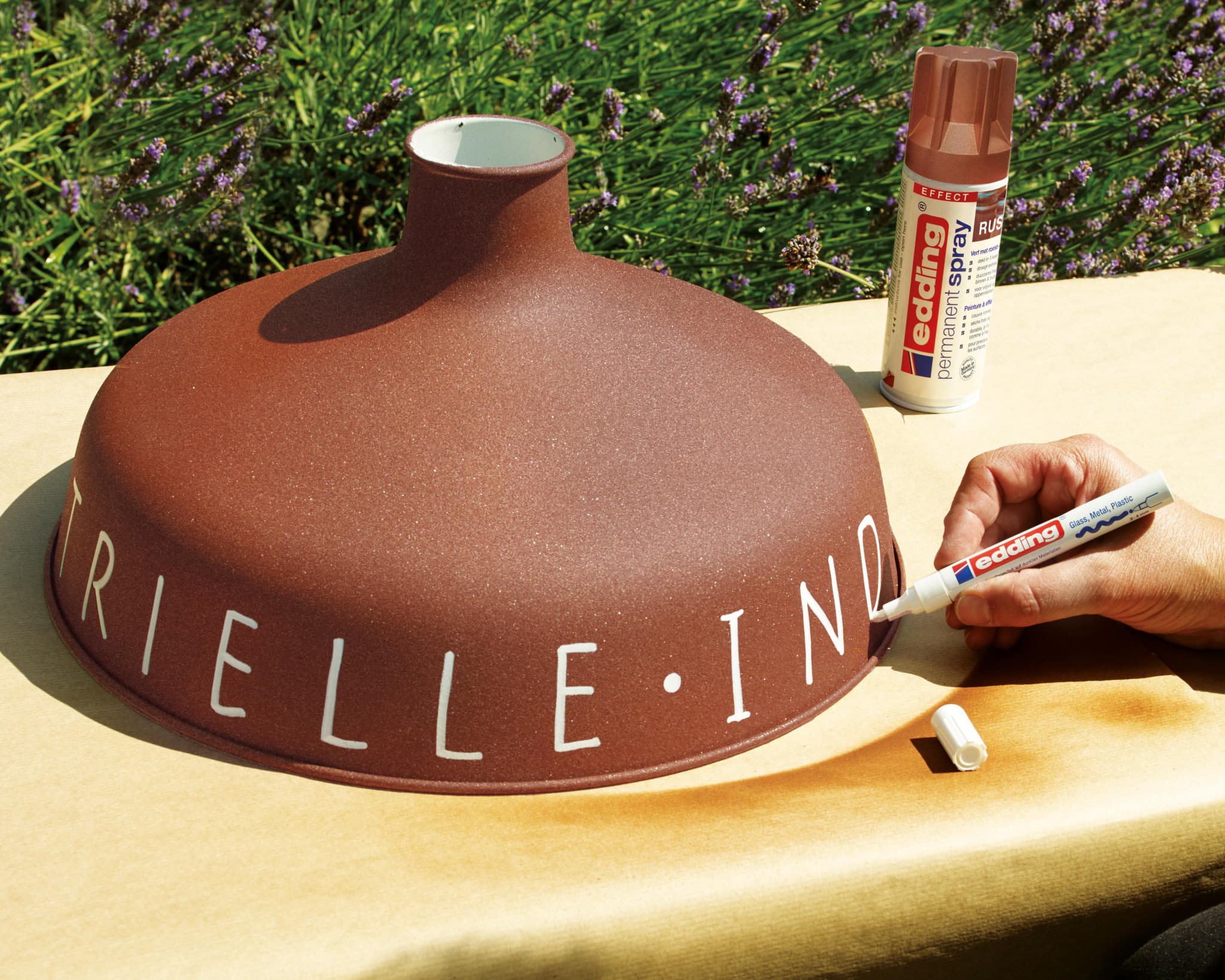 An einem sonnigen Tag im Freien einen Rost-Lampenschirm mit einem weißen Lackmarker individuell gestalten, mit einer Dose edding 5200 Permanentspray Effekt Acryllack in der Nähe.