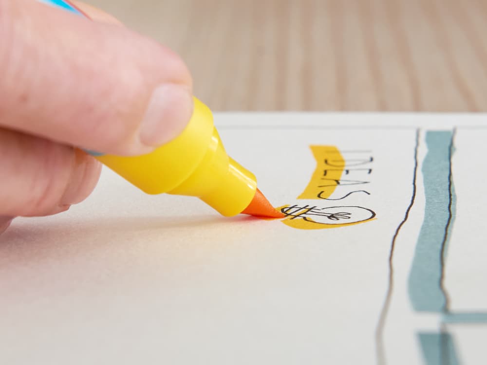 A hand holds an edding 385 Flipchart Effektmarker with a Pinselspitze, sketching a lightbulb next to "IDEAS" on lined paper featuring a light blue section.