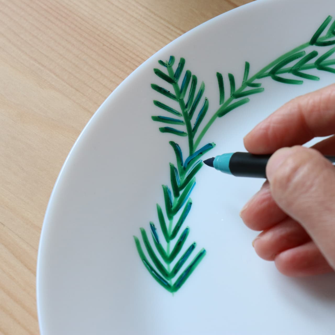 Mit dem edding 4200 Porzellanpinselstift von edding verziert eine Hand einen weißen Keramikteller mit einem Blattzweig-Motiv auf hellem Holzuntergrund. Das fertige Design ist spülmaschinenfest.