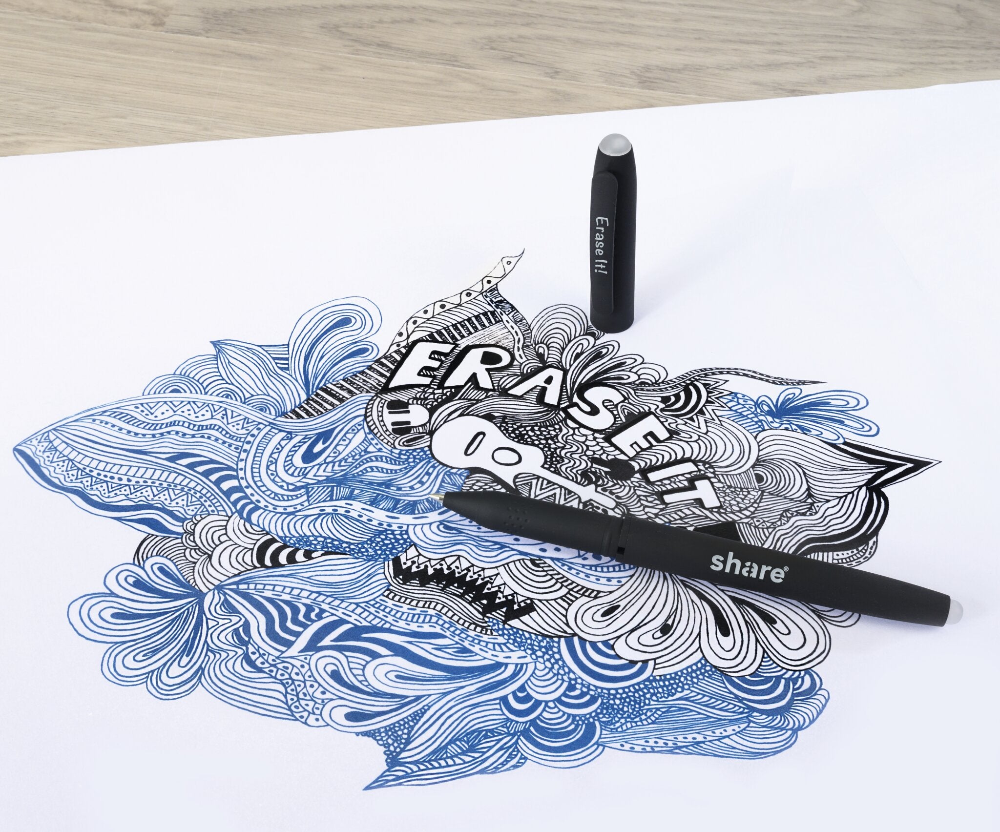 Ein detailliertes blaues und schwarzes abstraktes Doodle erscheint auf weißem Papier. Zwei share Gelroller M radierbar-Stifte aus dem share 2er-Set, einer mit der Aufschrift "Eraser" und einer mit der Aufschrift "Share", liegen auf dem Blatt, "ERASE IT" ist im Motiv sichtbar.