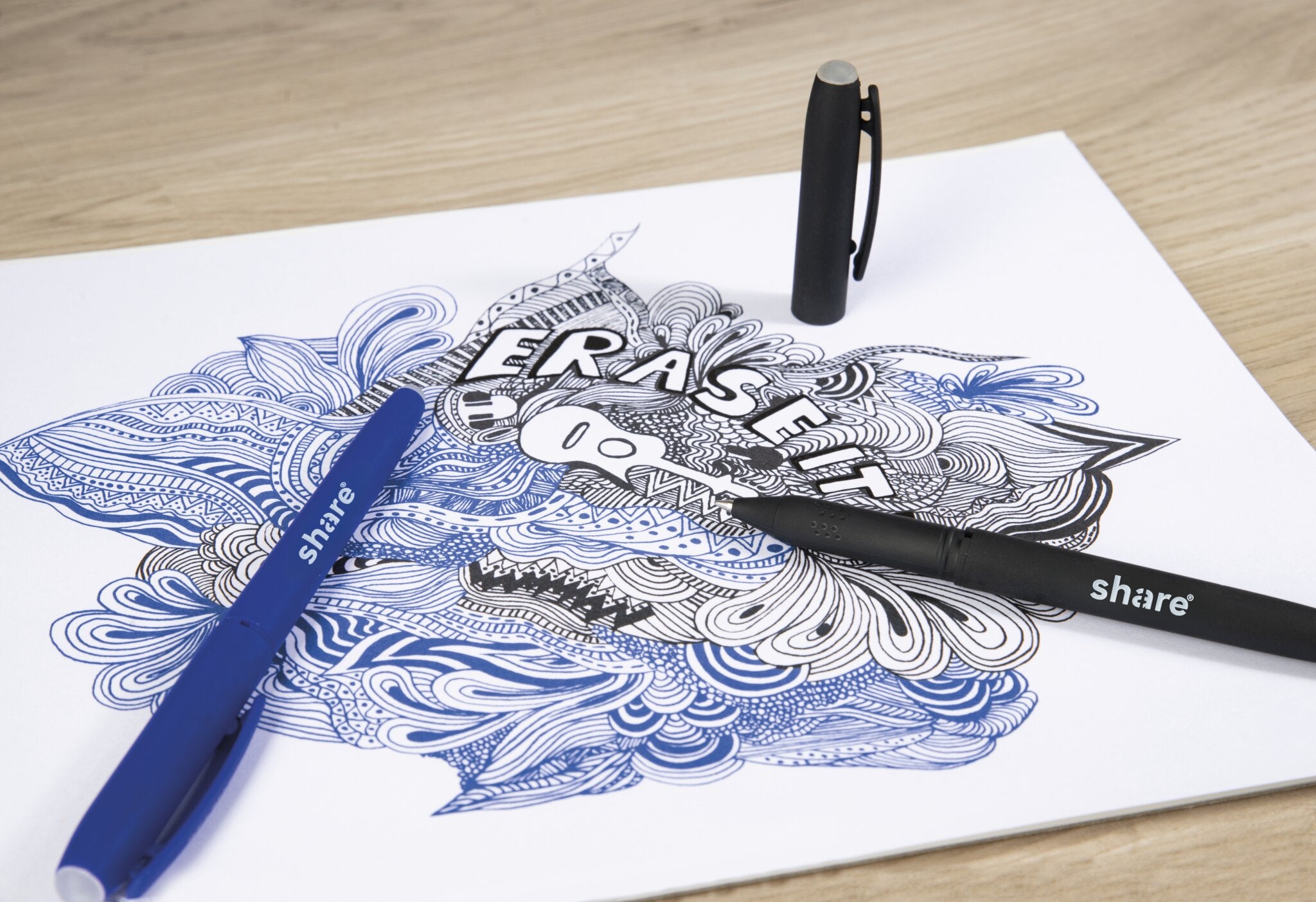 Ein detailliertes blaues und schwarzes Gekritzel auf weißem Papier mit dem Schriftzug "ERASE IT" in der Mitte. Zwei share Gelroller M radierbar Stifte aus dem 2er-Set von share, ein blauer und ein schwarzer, sowie eine Kappe, liegen auf dem Papier auf einer Holzunterlage.