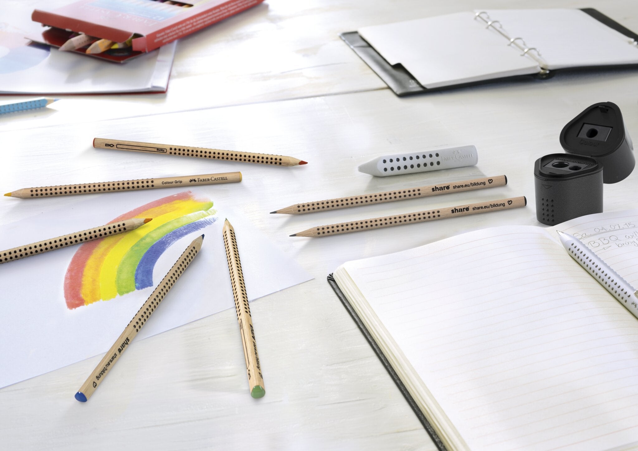 Ein weißer Schreibtisch mit Buntstiften, eine Anspitzer Dreifachdose, eine Regenbogenzeichnung, ein aufgeschlagenes Notizbuch, ein Federmäppchen aus recyceltem Kunststoff und eine Mappe mit losem Papier für nachhaltigen Konsum.