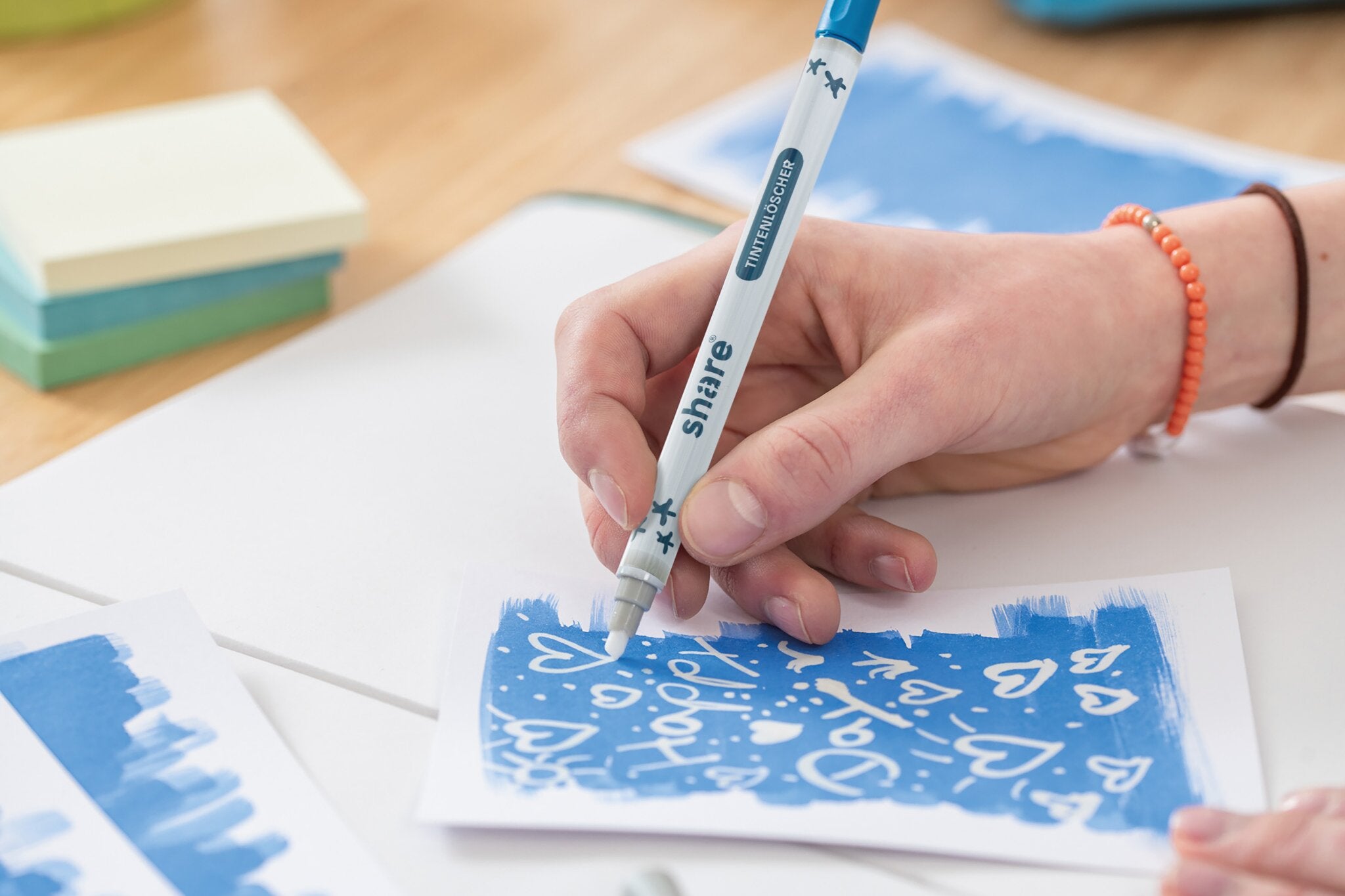 Eine Hand benutzt das share Tintenlöscher 2er-Set von share aus recyceltem Kunststoff, um "Happy" zu schreiben und Herzen auf eine blau-weiße Karte zu zeichnen. Haftnotizen und andere Karten liegen daneben auf dem Holztisch.