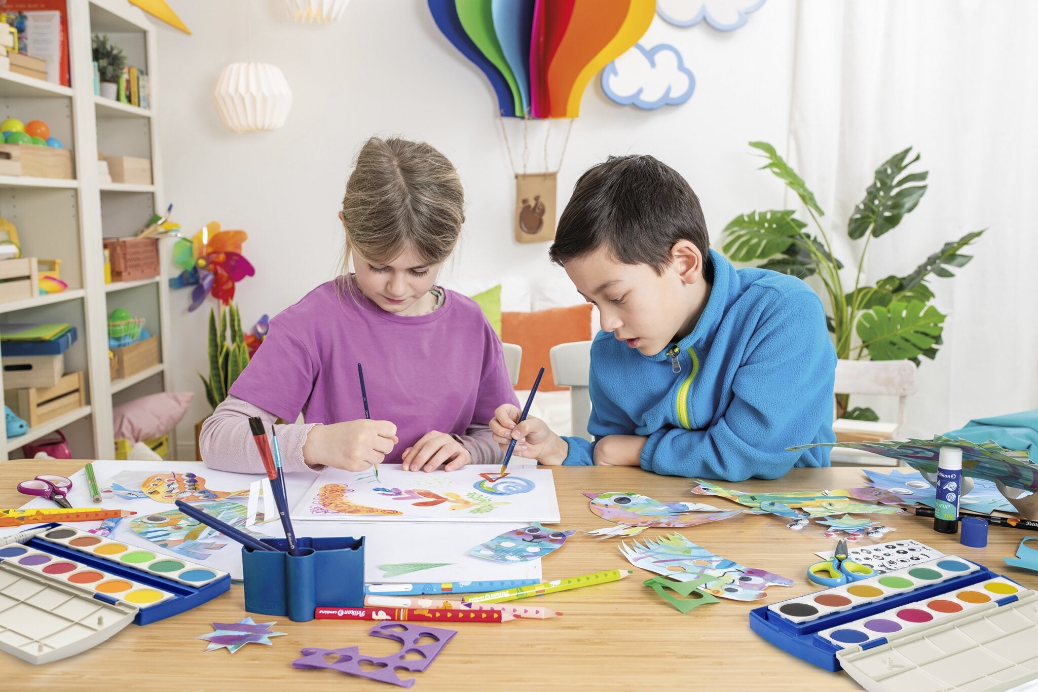 Zwei Kinder teilen sich den Deckfarbenkasten 12 Farben von share und malen mit Wasserfarben und Pinseln bunte Fische und Formen. Der Tisch ist mit Malutensilien, Papier und ihren Kunstwerken gefüllt.