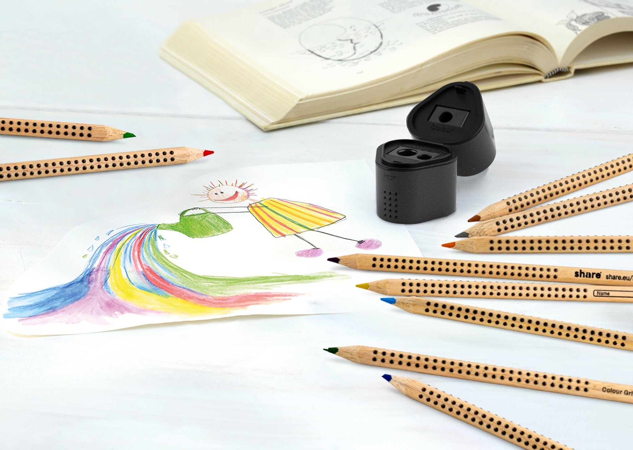 Eine bunte Kinderzeichnung mit einer lächelnden Person und einem Regenbogen liegt auf einem Tisch, umgeben von Buntstiften, der Anspitzer Dreifachdose von share und einem aufgeschlagenen Schulbuch im Hintergrund.