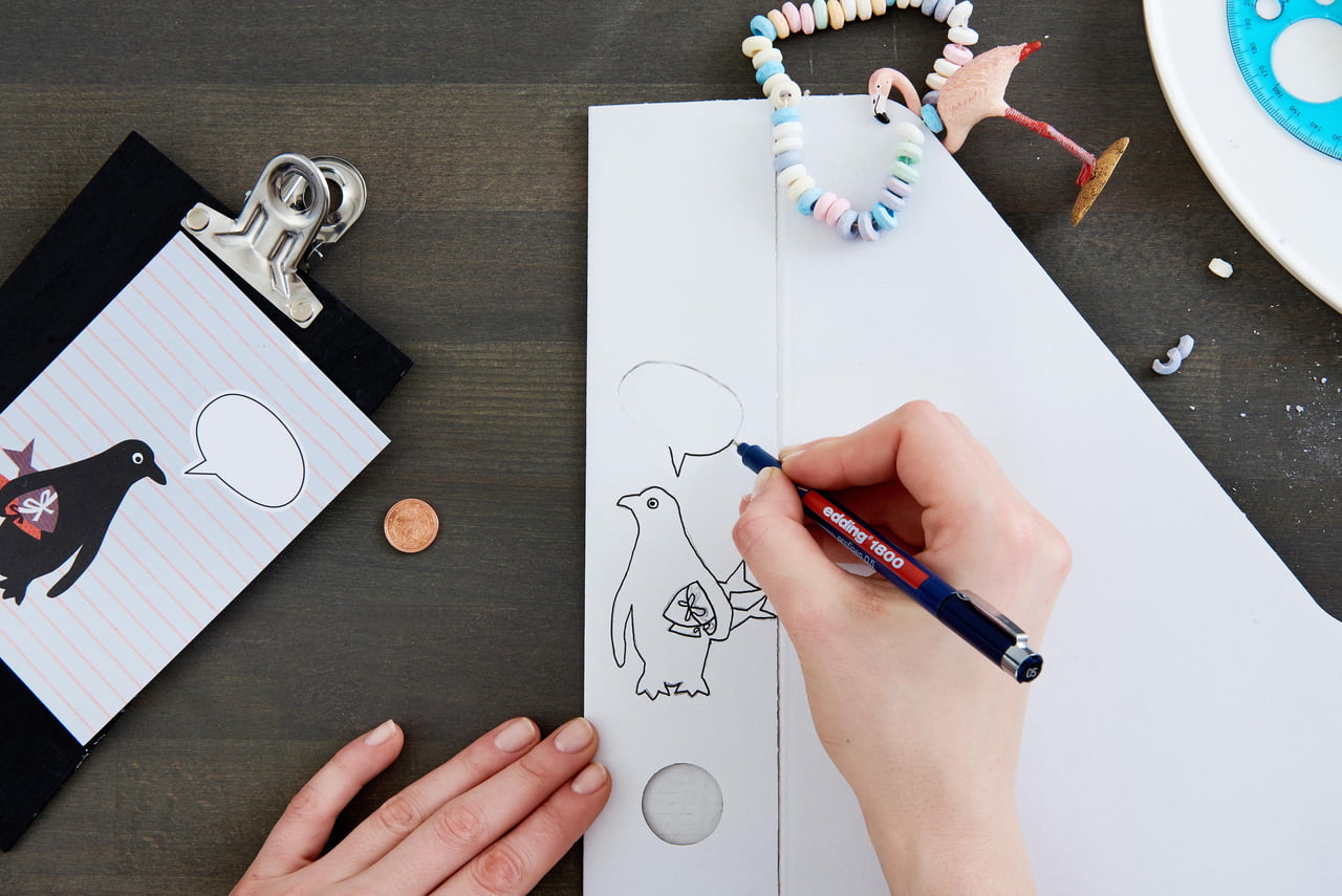 Hände skizzieren einen Cartoon-Pinguin mit Sprechblase auf weißem Papier, daneben kreatives Zubehör und edding 1800 Präzisionsfeinschreiber-Kritzeleien auf einer dunklen Schreibtischoberfläche.