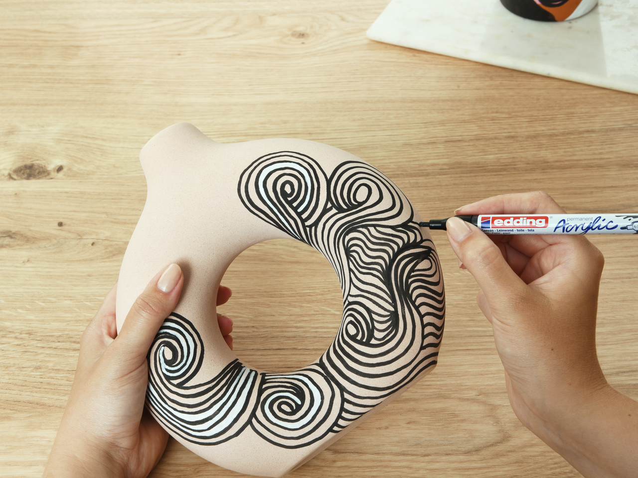 Handgezeichnete Kunst in Arbeit: Eine Person verziert kreativ ein beiges Nackenkissen mit einem schwarzen Wirbelmuster mit einem edding 5300 Acrylmarker fein.