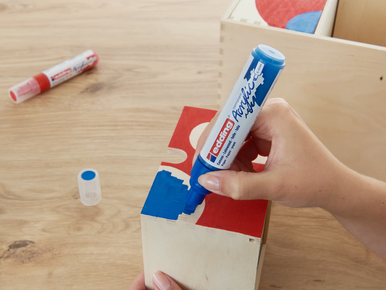 Eine Person trägt während eines Bastelprojekts einen blauen edding 5000 Acrylmarker auf ein rotes Kartonstück auf.