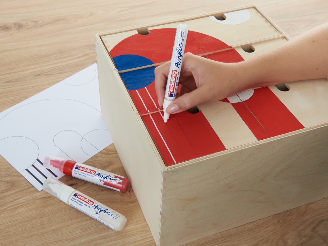 Handzeichnen eines roten und blauen Designs auf einem Cornhole-Holzbrett mit edding 5100 Acrylmarker medium 5er-Set, mit zusätzlichen Farbmarkern und einer daneben liegenden Skizze.