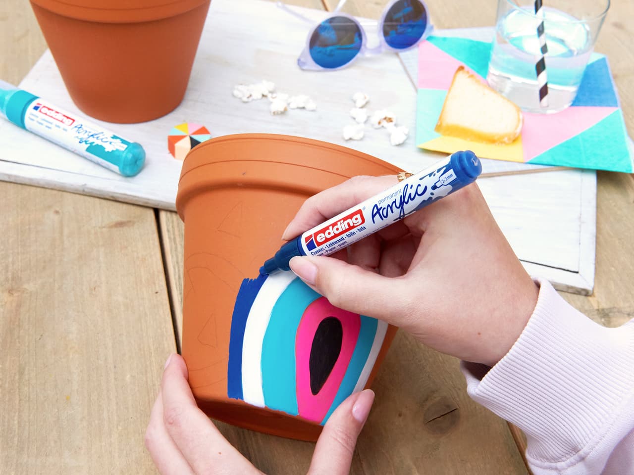 Eine kreative Hand, die mit edding 5100 Acrylmarker medium 5er-Set farbenfrohe abstrakte Designs auf einen Terrakotta-Blumentopf malt, umgeben von Künstleraustattung und einer entspannten Bastelatmosphäre.