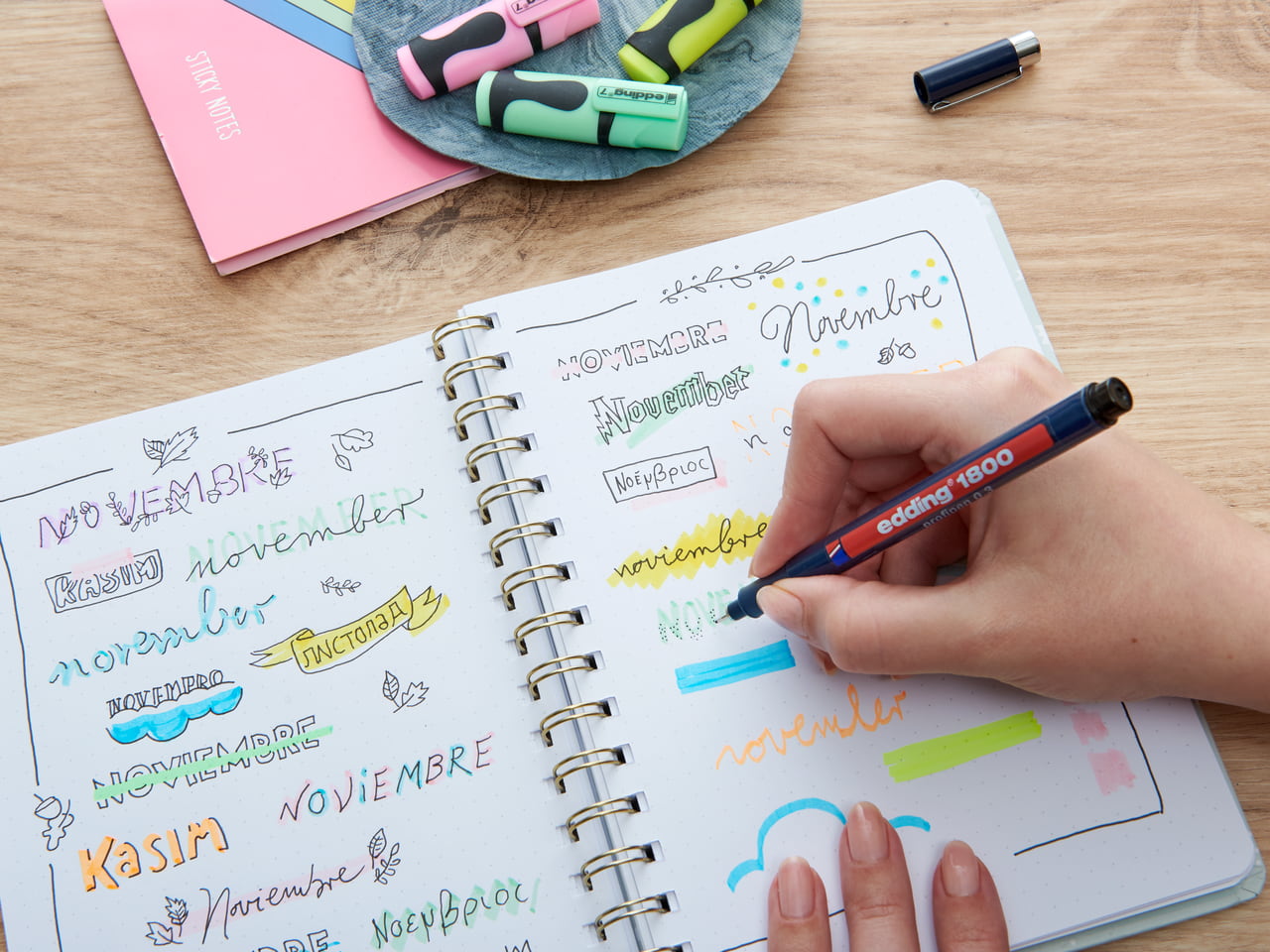 Eine Person übt das Handlettering des Wortes „November“ in verschiedenen Stilen und Sprachen in einem Notizbuch, umgeben von Edding-Markern und Schreibwaren.