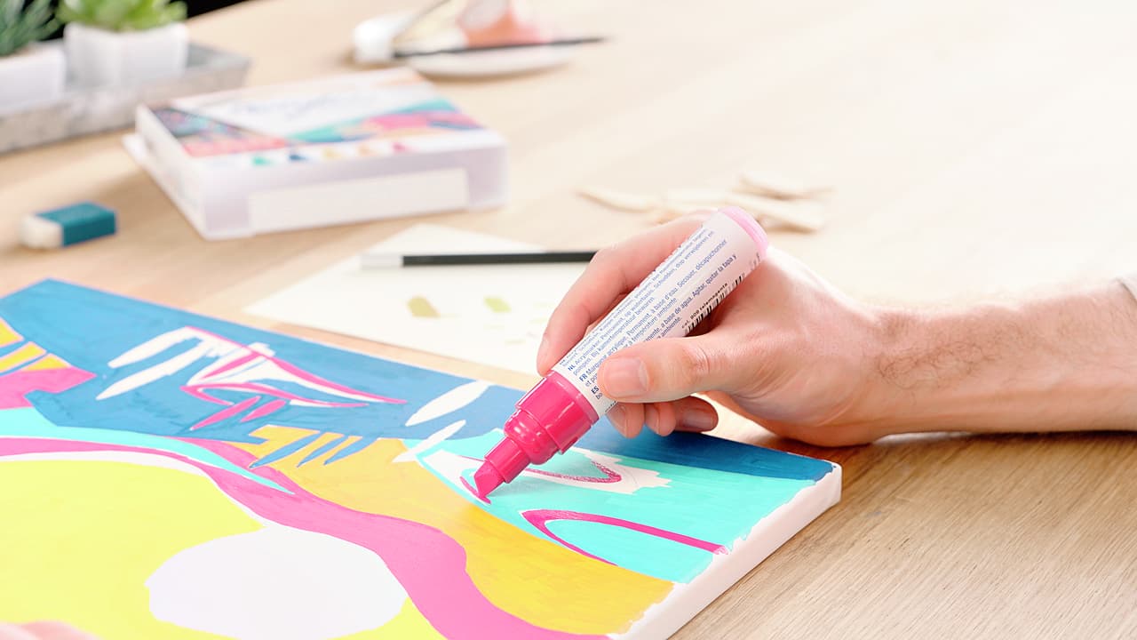 Die Hand einer Person hält ein rosa edding 5000 Acrylmarker breit 5er-Set und fügt Details zu einem farbenfrohen abstrakten Gemälde auf einem Holztisch hinzu.