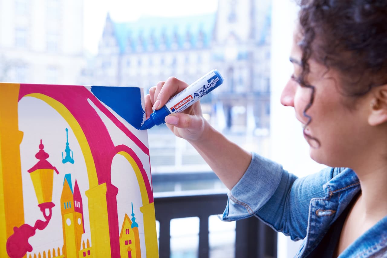Ein Künstler, der mit einem blauen edding 5000 Acrylmarker Details zu einem farbenfrohen Stadtgemälde hinzufügt.
