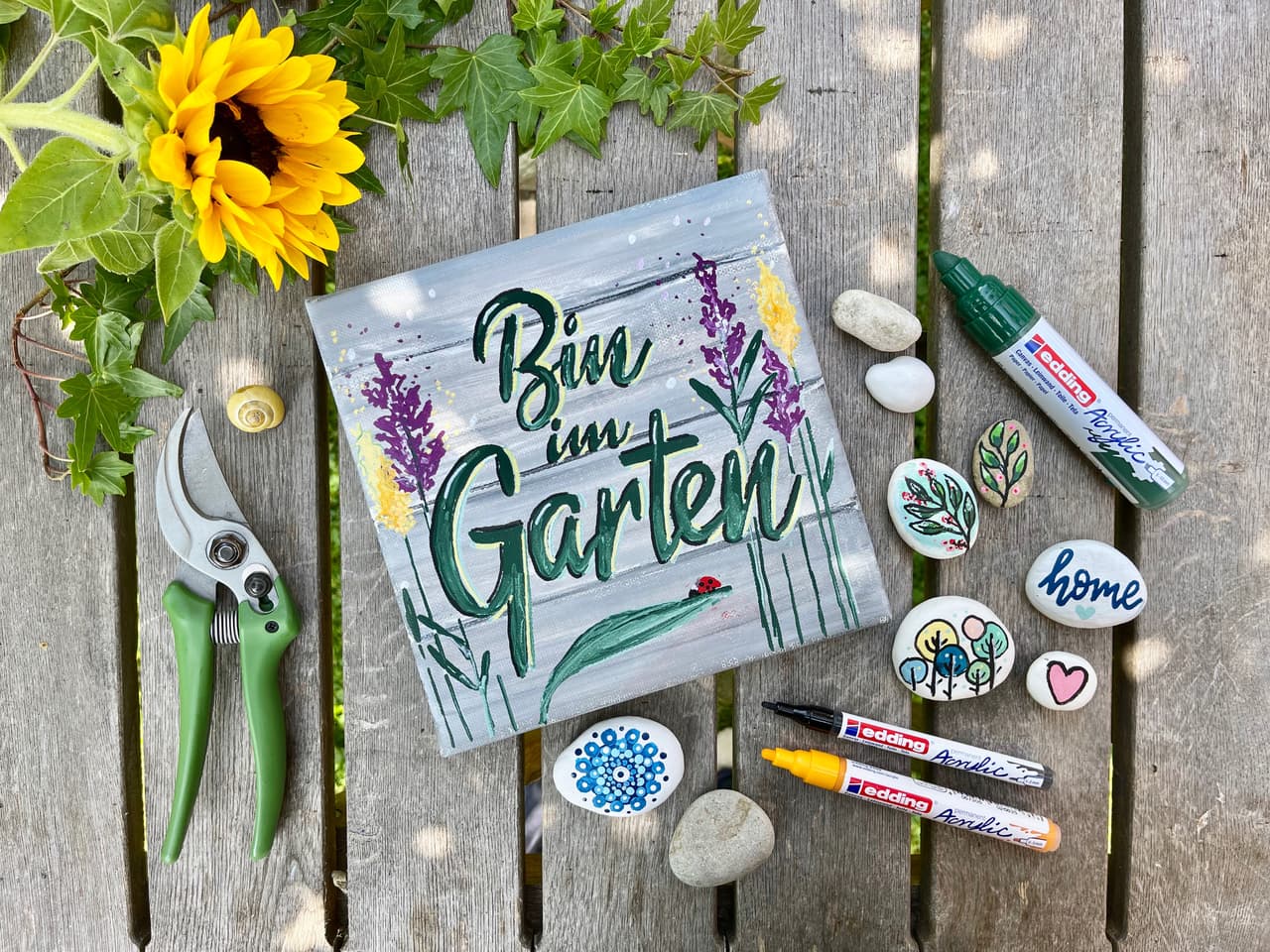 Eine fröhliche und kreative Bastelszene zum Thema Garten mit einem handgemalten edding 5000 Acrylmarker breit 5er-Set-Schild mit der Aufschrift „bin im garten“, umgeben von bemalten Steinen und einer Gartenschere.