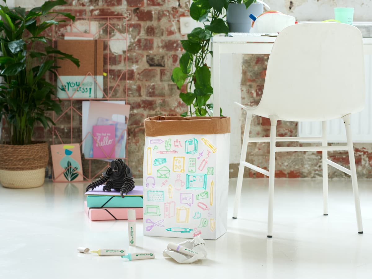 Kreativer Arbeitsbereich mit farbenfroher Papeterie, darunter edding 24 EcoLine Textmarker, einem weißen Stuhl und einer Pflanze, der eine organisierte und dennoch lebendige Atmosphäre für Produktivität und Inspiration bietet.