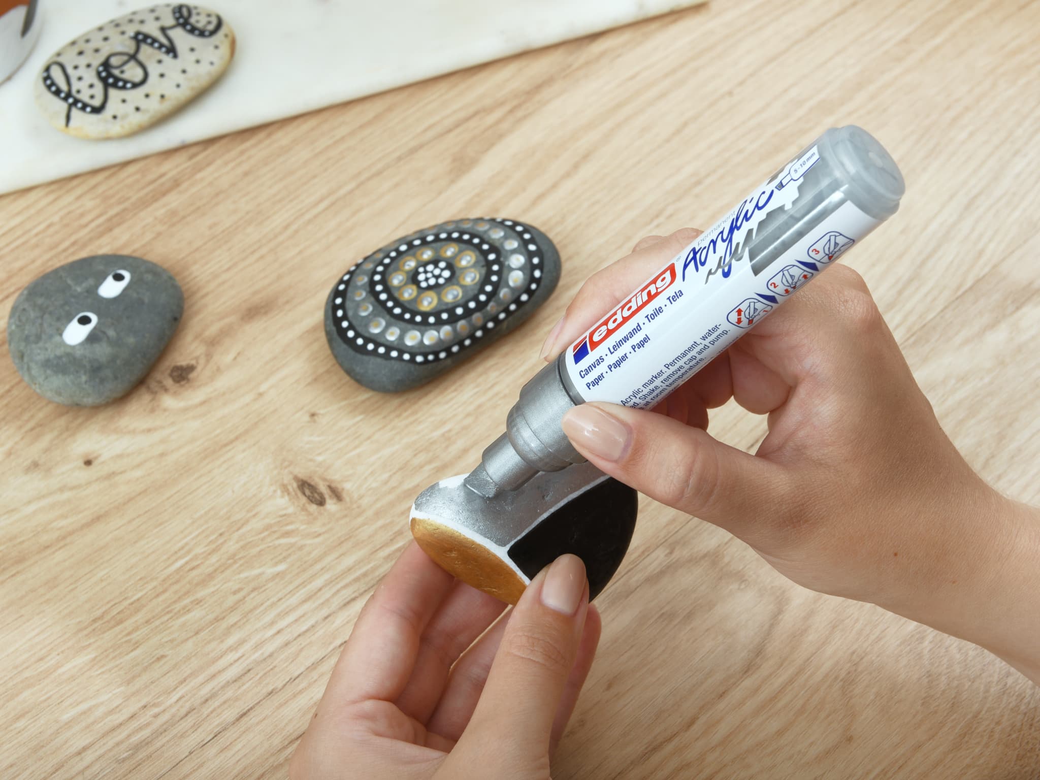 Eine Person trägt Klebstoff aus einer Tube auf einen glatten Stein auf, vermutlich für ein Bastelprojekt, mit verzierten Steinen im Hintergrund mit hochpigmentierter Tinte und edding 5000 Acrylmarker breit.