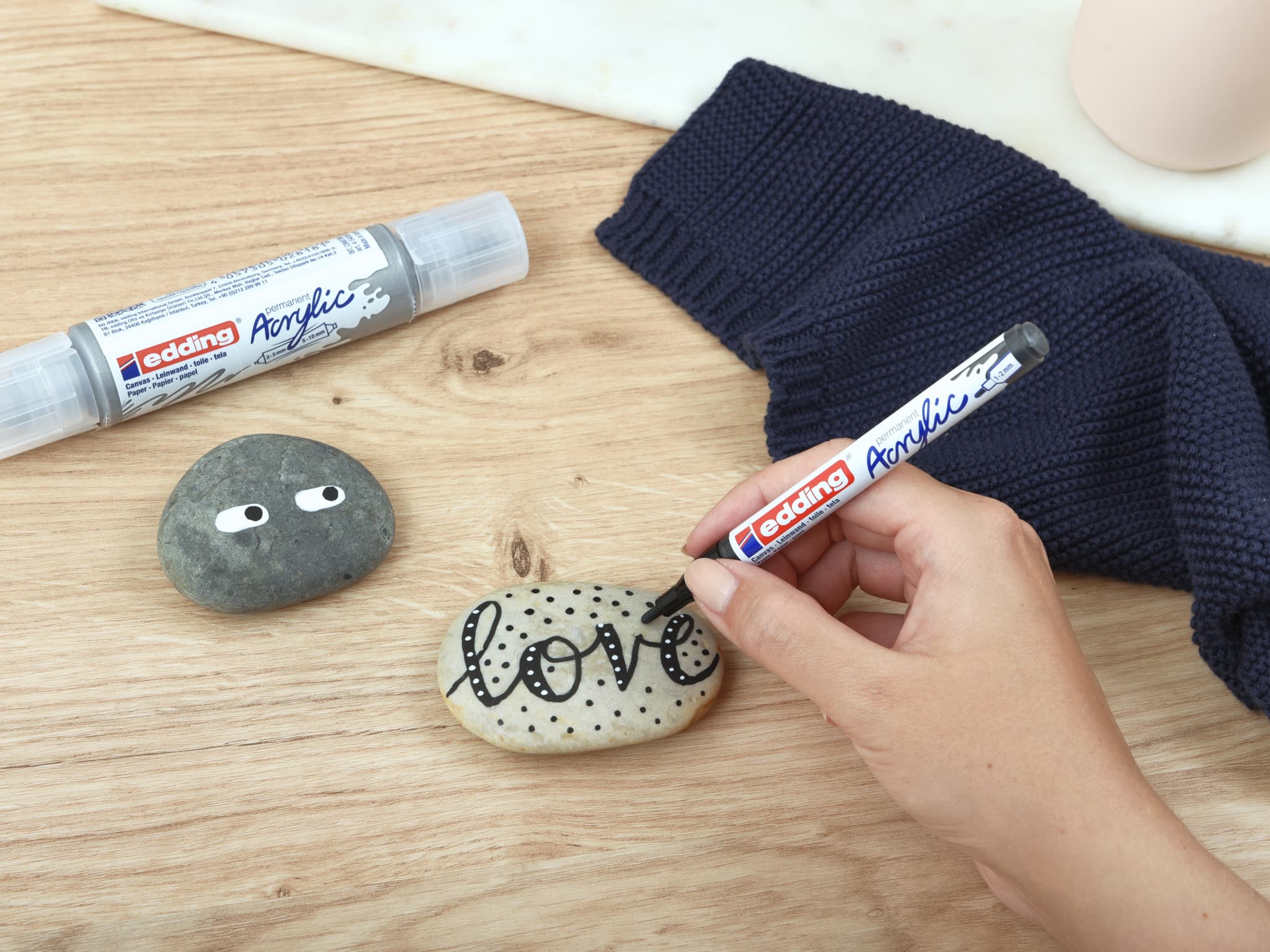 Die Hand einer Person verziert mit einem edding 5300 Acrylmarker fein einen glatten Stein mit dem Wort „Love“, während auf einer Holzoberfläche ein weiterer dekorierter Stein und Malutensilien sichtbar sind.