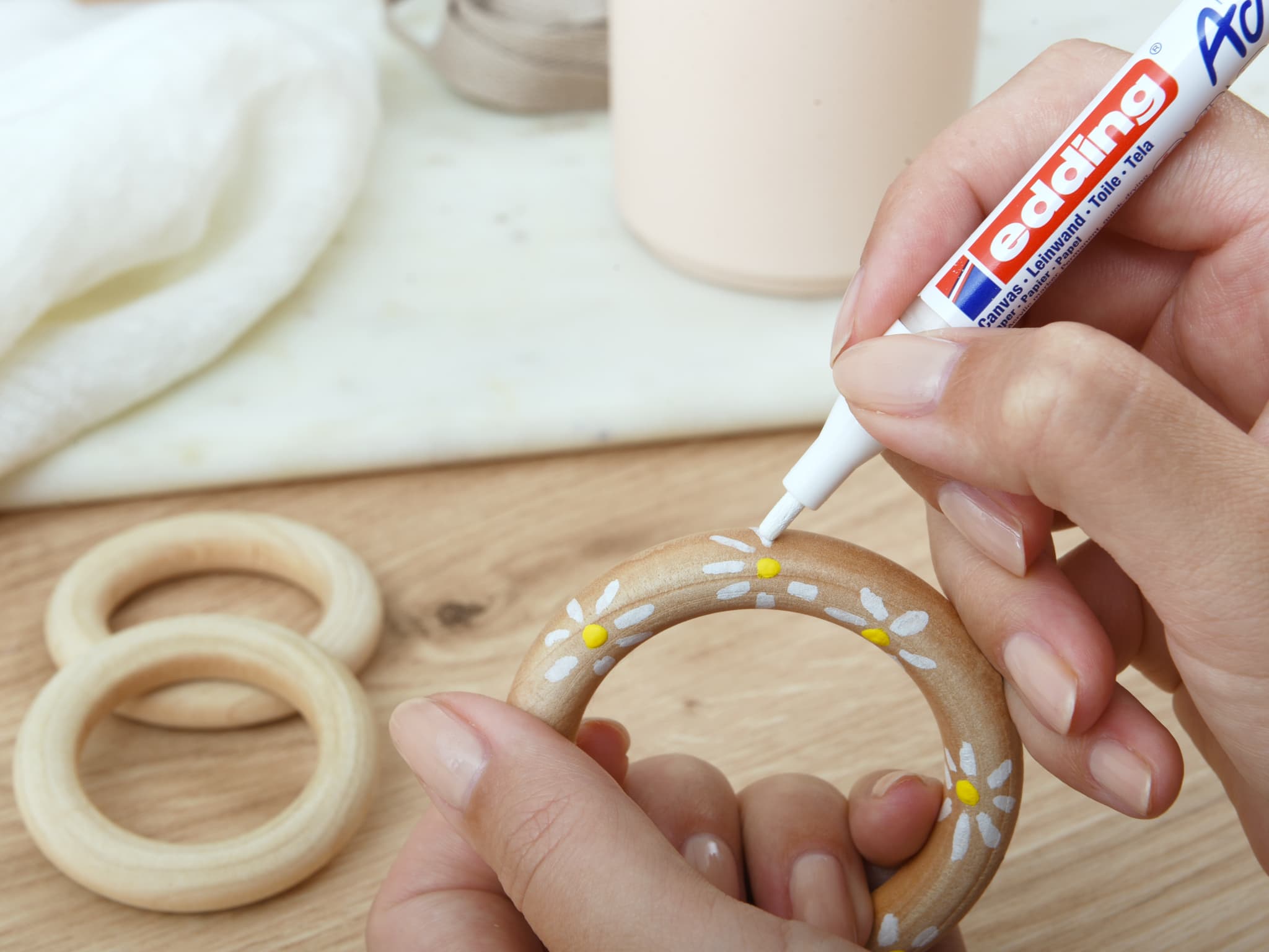 Bastelzeit: Hände verzieren einen Holzring mit weißem edding 5300 Acrylmarker fein und fügen zarte Gänseblümchenmuster für eine hausgemachte Note hinzu.