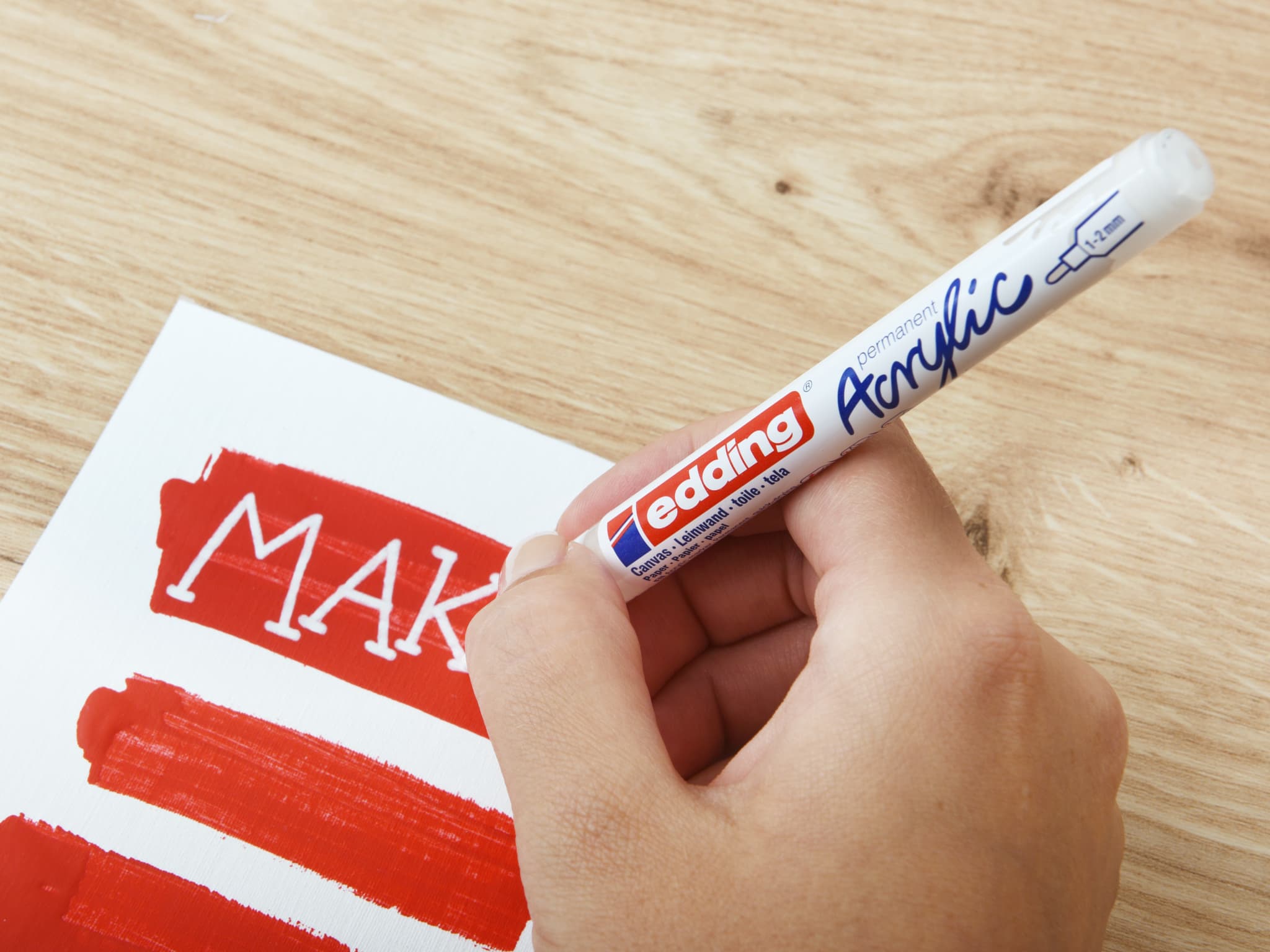 Eine Person schreibt mit einem edding Marker das Wort „make“ in rotem edding 5300 Acrylmarker fein auf ein weißes Blatt Papier.