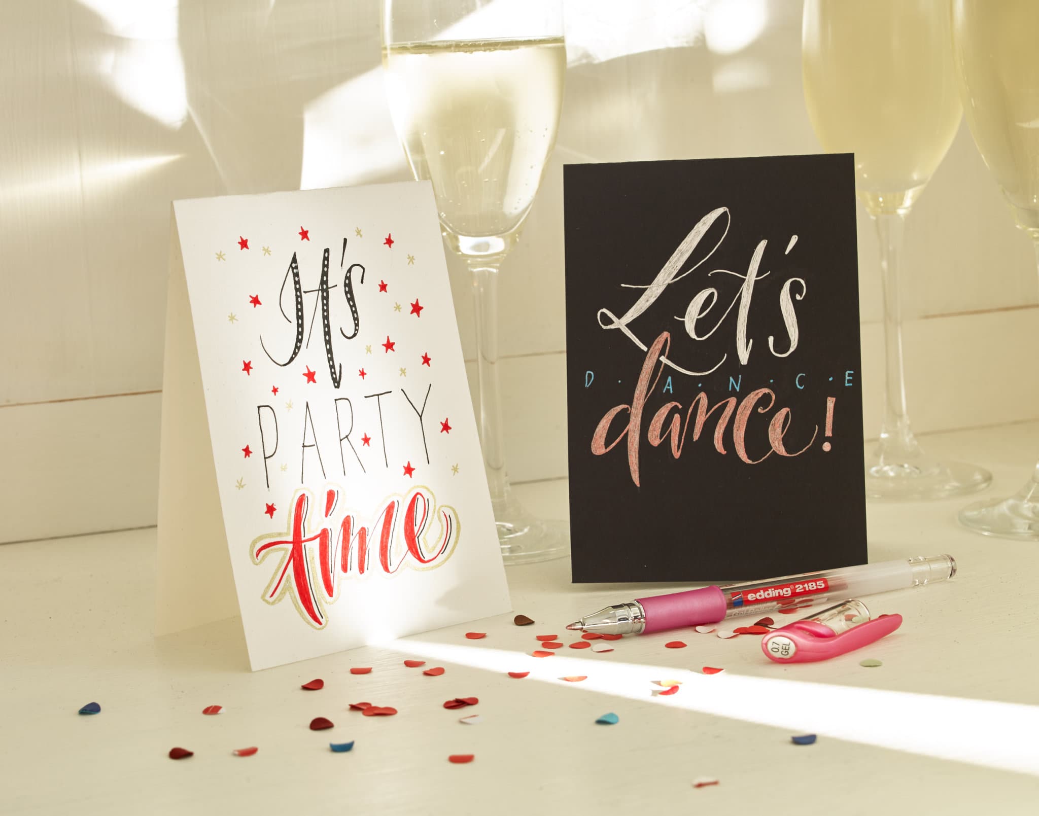 Zwei festliche Karten auf einem Tisch, eine mit der Aufschrift „Es ist Partyzeit“ und die andere „Lass uns tanzen“, mit herumstreuendem Konfetti und einem Champagnerglas, das für festliche Stimmung sorgt. Das edding 2185 Gelroller 5er-Set ist perfekt für jeden Anlass.