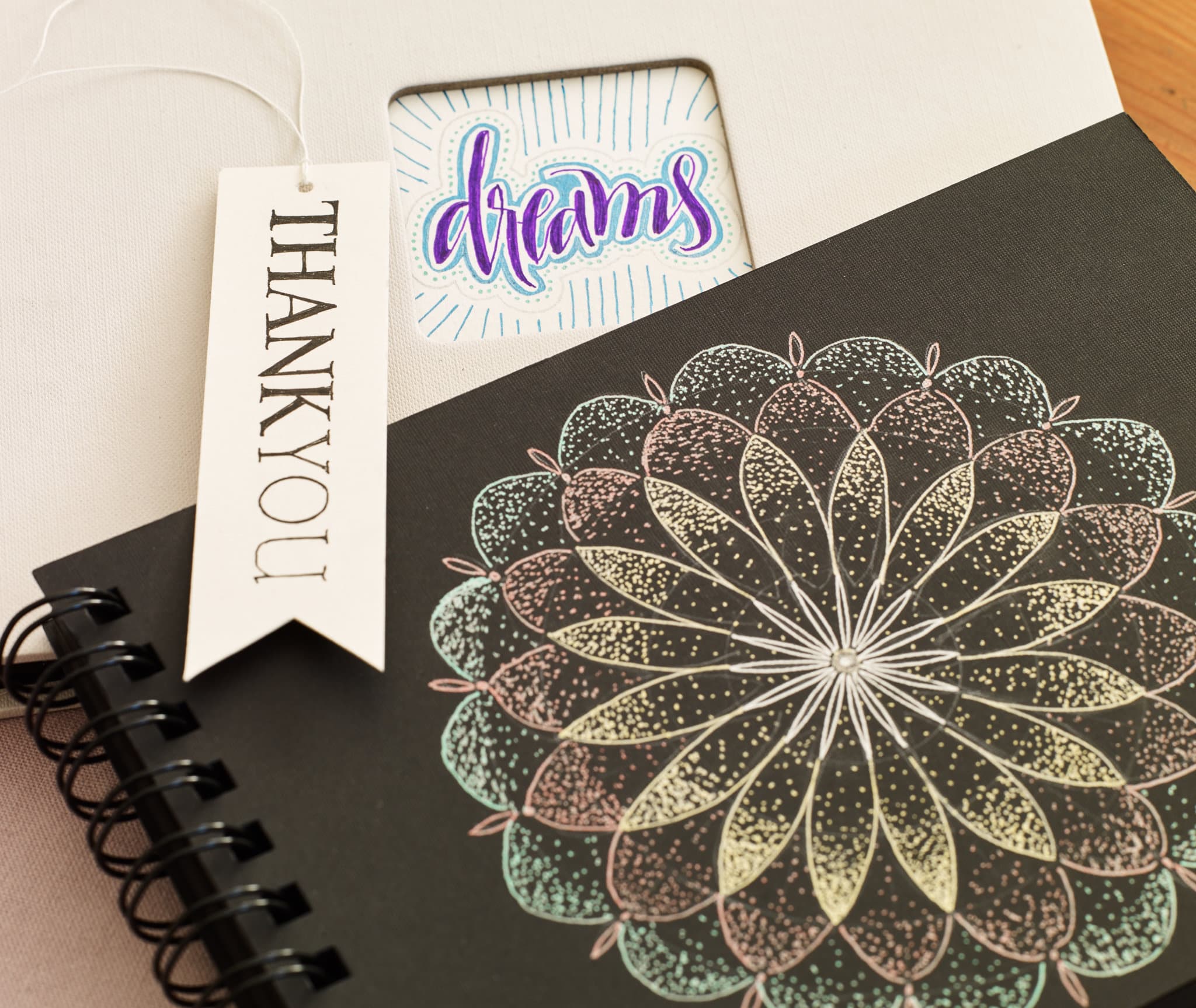 Ein kunstvolles schwarzes Notizbuch mit einem aufwendigen Mandala-Design in Metallic-Farben auf dem Einband, gepaart mit Tags mit der Aufschrift „Danke“ und „Träume“, die Gefühle der Dankbarkeit und Inspiration hervorrufen, von edding 2185 Gelroller.