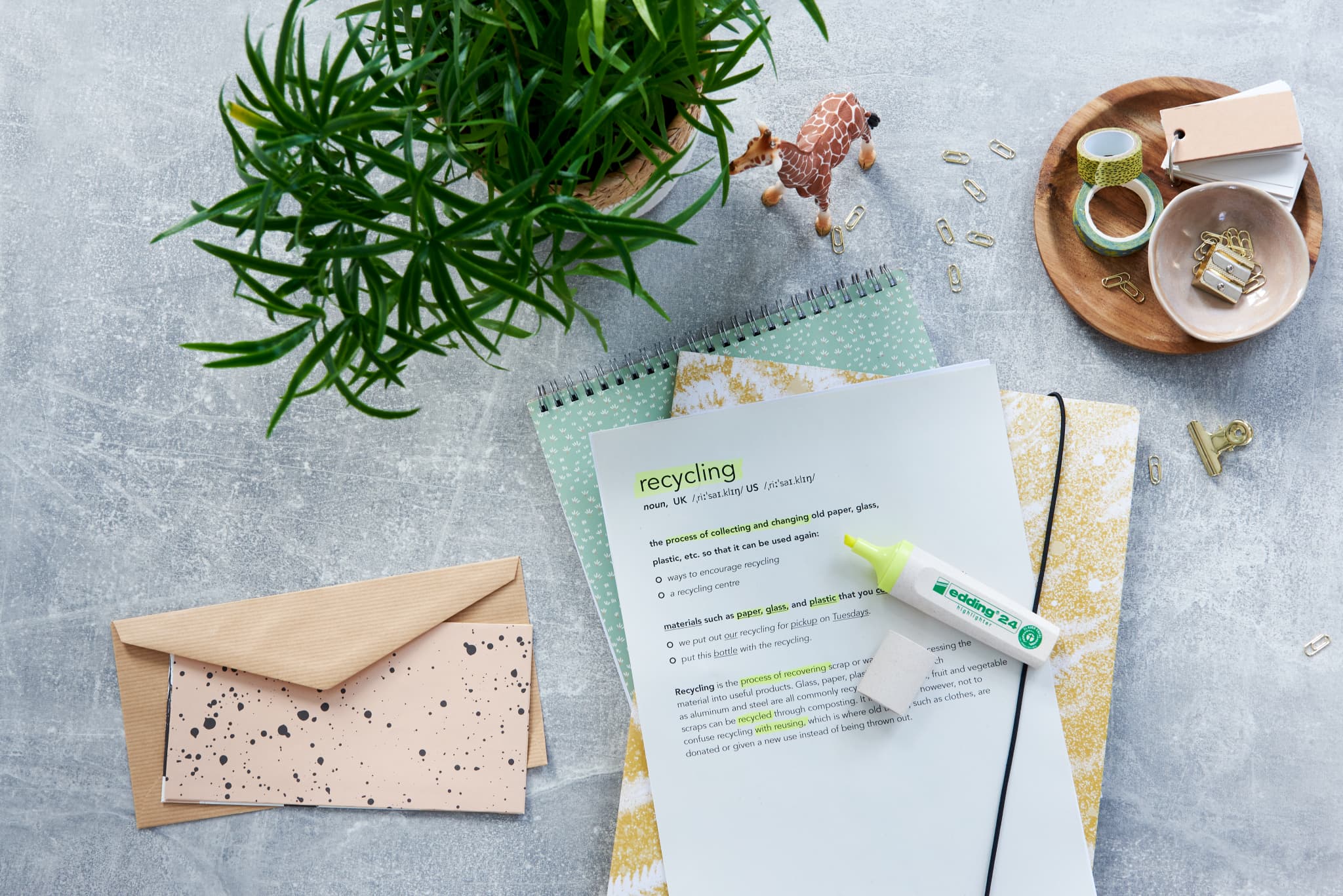 Eine flache Komposition mit umweltfreundlichen Büromaterialien, darunter ein Recycling-Tippblatt, eine Pflanze, ein Notizblock, Umschläge und ein edding 24 EcoLine Textmarker, alles auf einer strukturierten Oberfläche angeordnet.