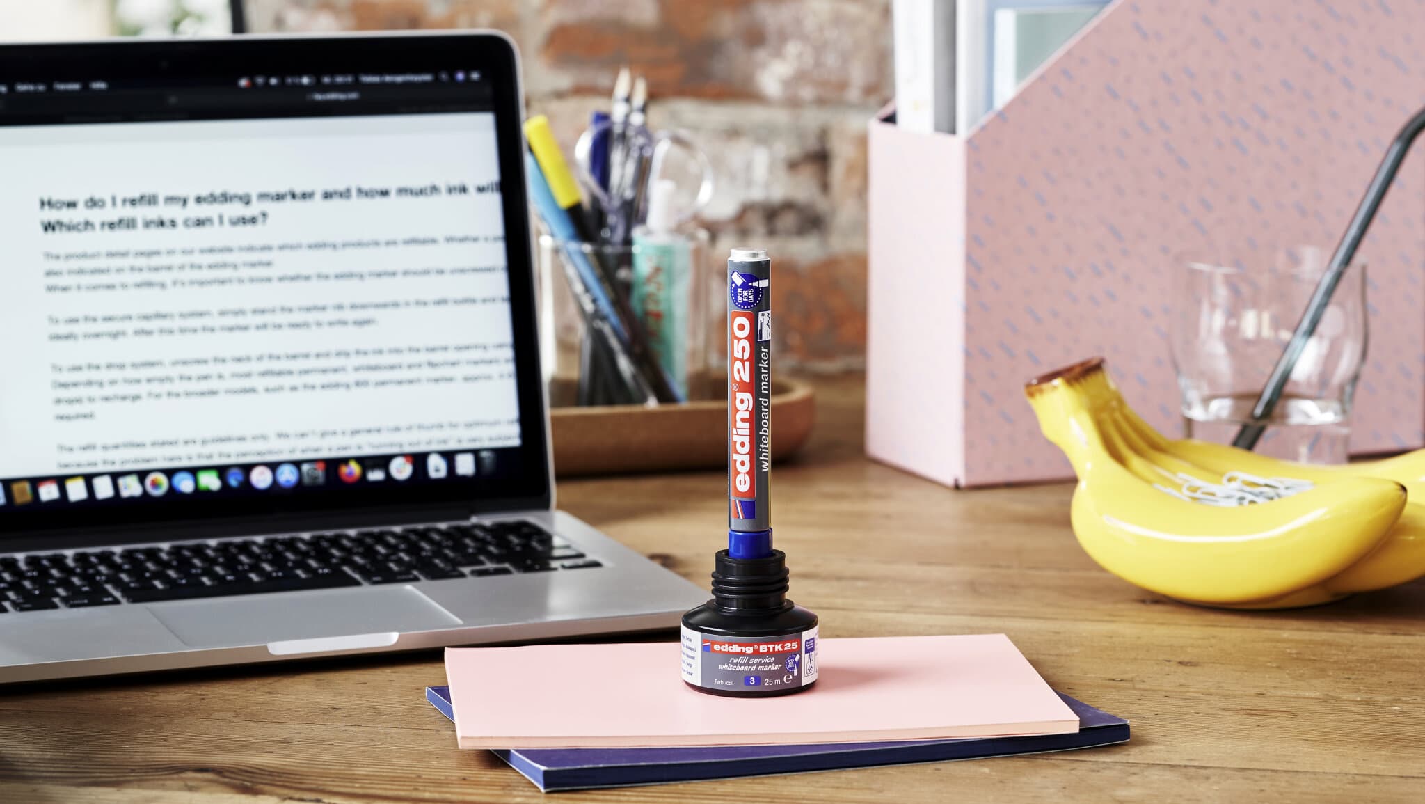 Ein ordentlicher Arbeitsplatz mit einem offenen Laptop, auf dem Text angezeigt wird, einer Flasche Kleber auf bunten Notizbüchern, einem bananenförmigen Telefonhalter und einer Brille daneben, daneben ein edding 250 Whiteboardmarker 4er-Set.