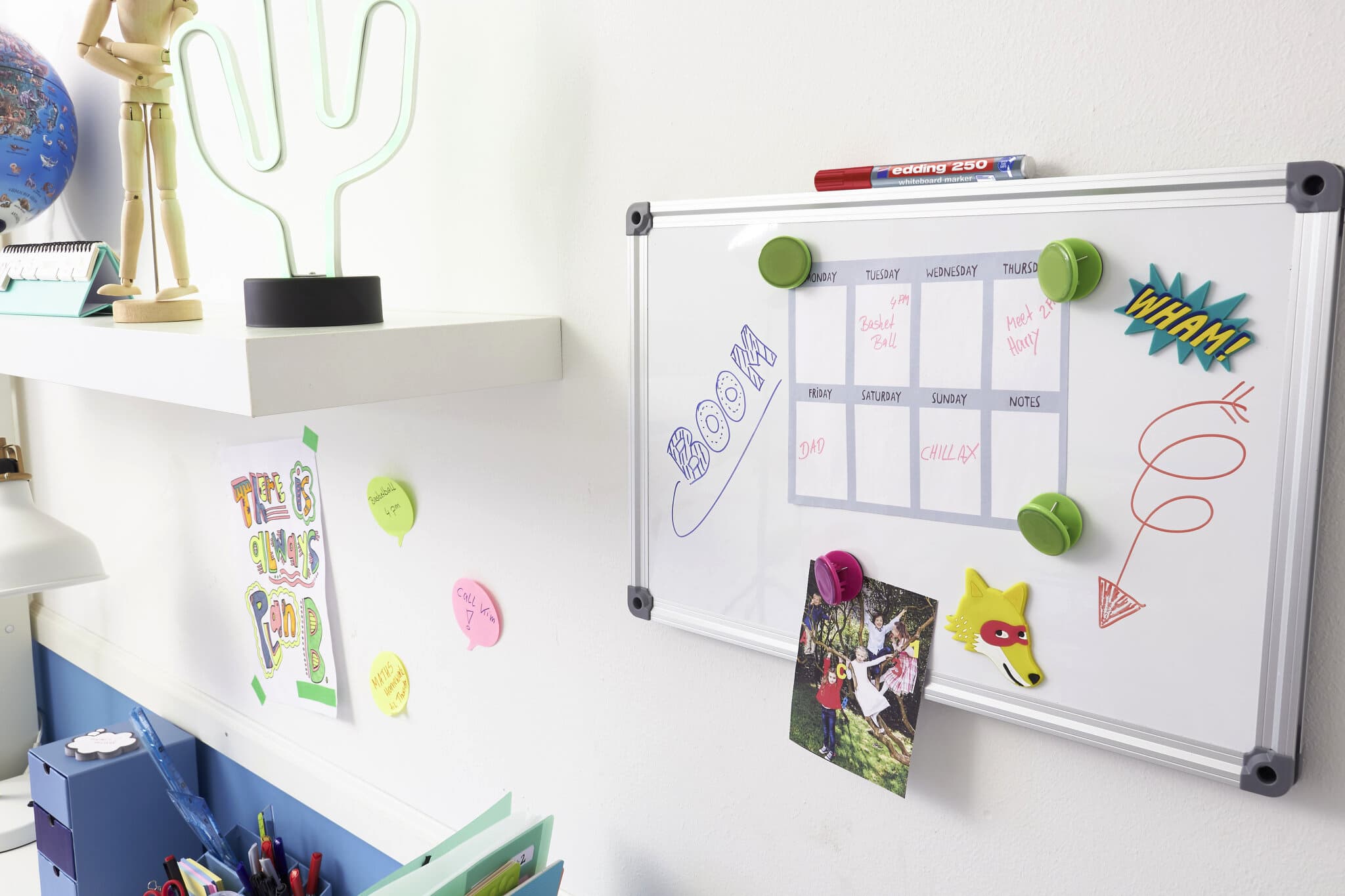 Eine lebendige und kreative Arbeitsplatzecke mit einem edding 250 Whiteboardmarker-Kalender, verziert mit bunten Magneten, Comic-Ausrufen und einem verspielten Regaldisplay mit einer Kaktuslampe und skurrilen Ornamenten. Ein Satz edding 250 Whiteboardmarker.