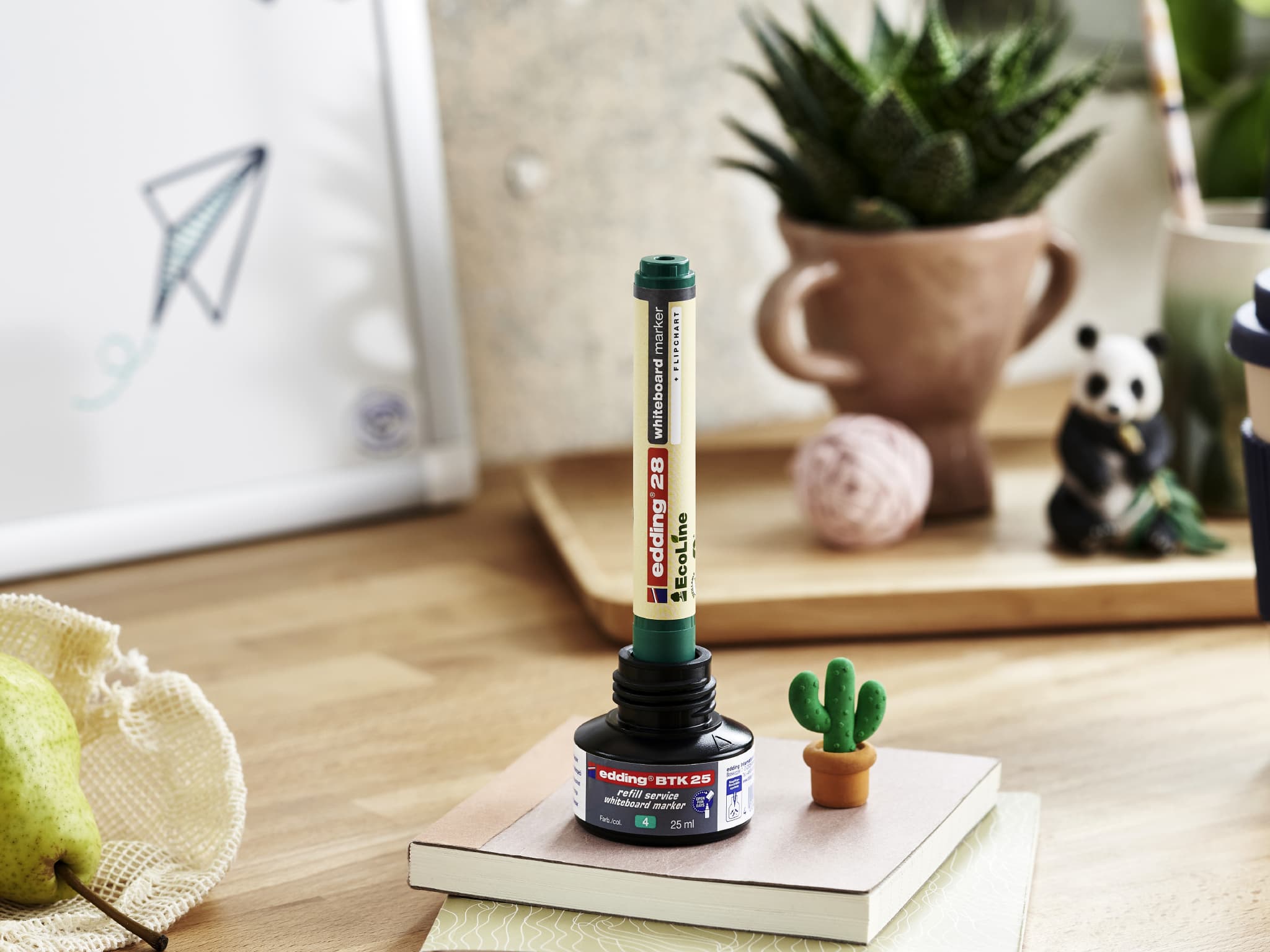 Ein kreatives Arrangement auf einem Holzschreibtisch mit einem Stapel farbiger Marker, balanciert auf einem kleinen Behälter aus recyceltem Material, mit Büro- und Dekorationsgegenständen, darunter ein edding 29 EcoLine Whiteboardmarker.